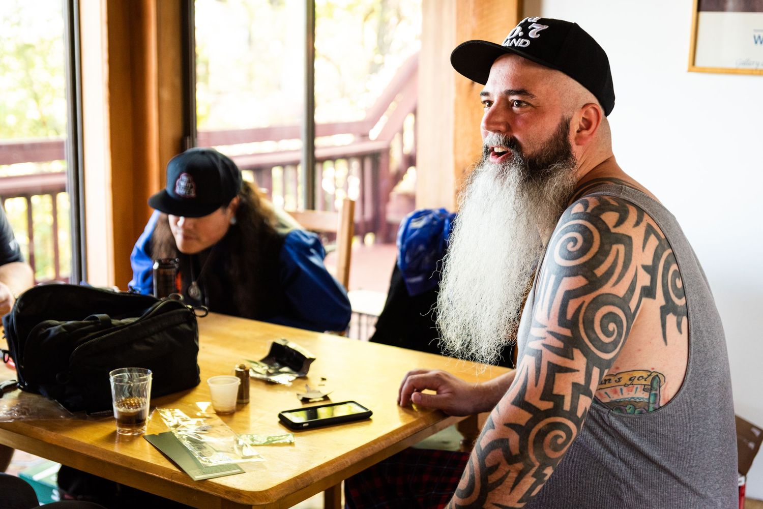 Two men sit at a table, with one sporting distinctive tribal arm tattoos and a long white beard.
