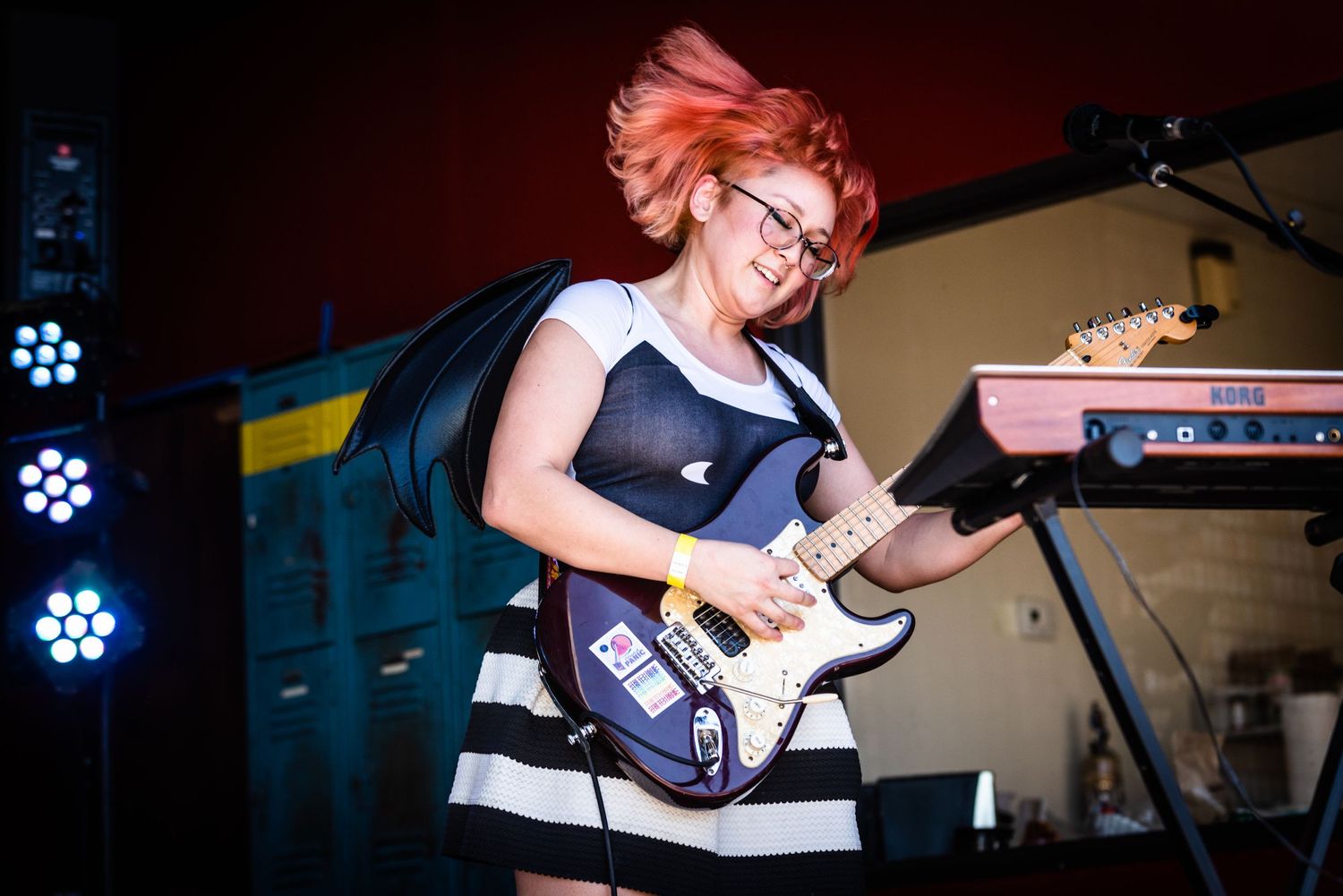 Musician in striped dress performs on electric guitar with colorful stage lighting.