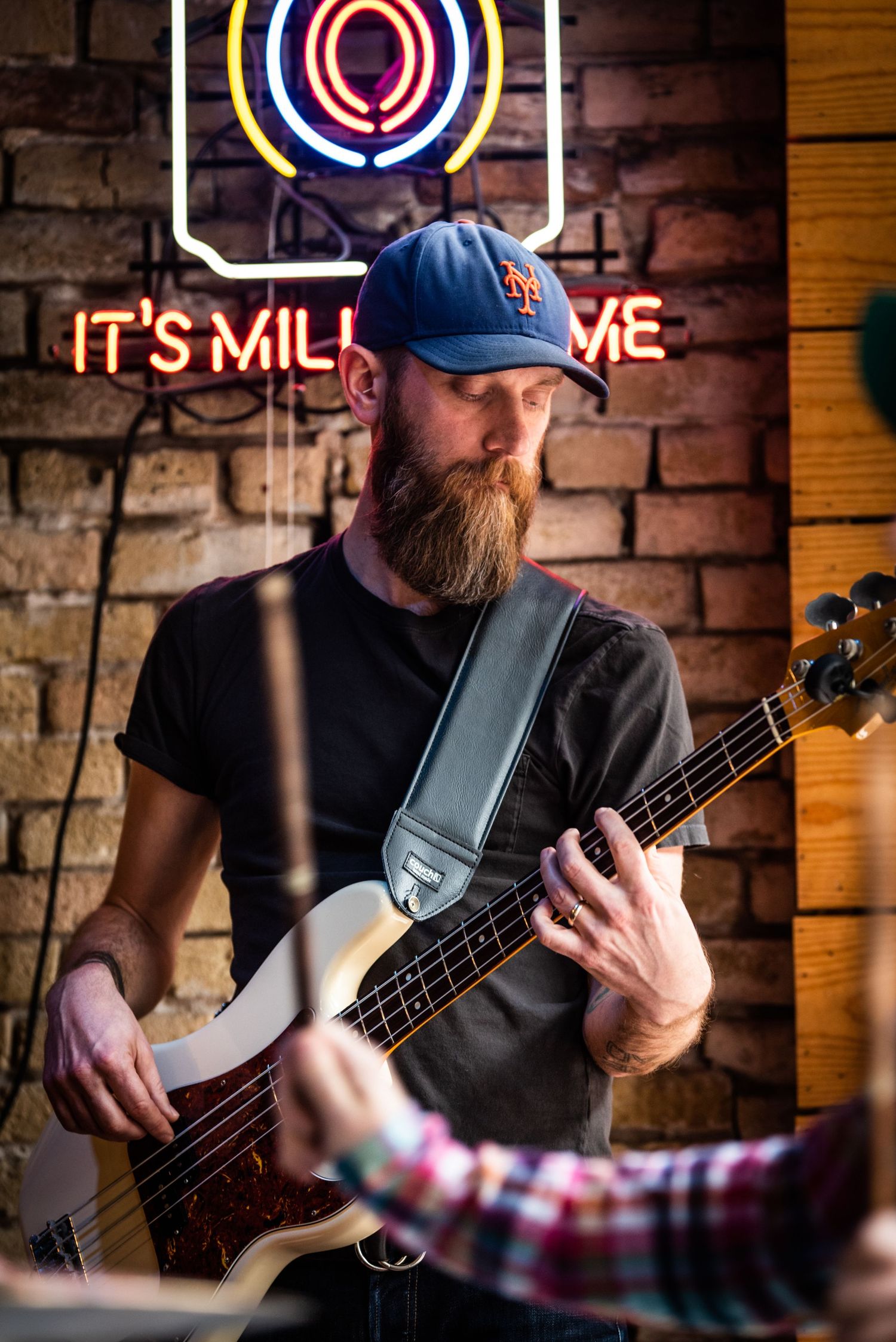 Bass guitarist in baseball cap performing beneath colorful neon signage at brick-walled venue.