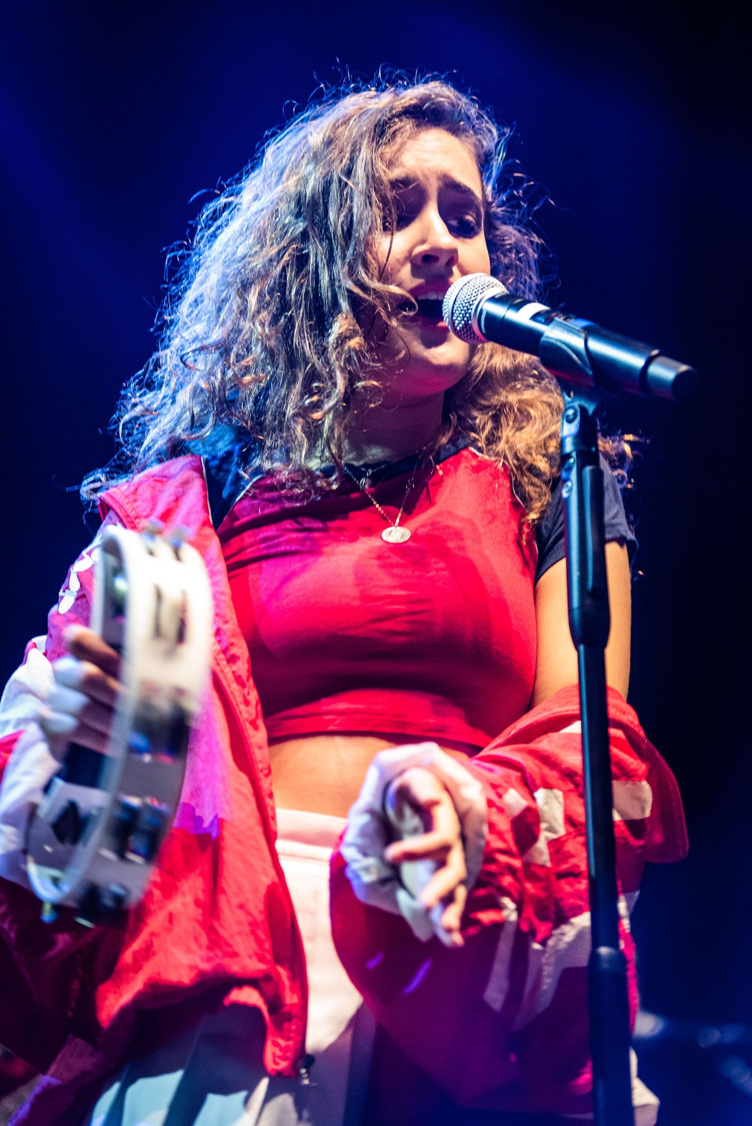 Performer in red crop top and jacket sings into microphone with blue stage lighting.