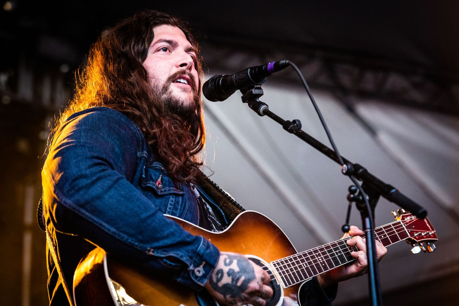 Musician with long dark hair performs on acoustic guitar under stage lighting wearing denim jacket.