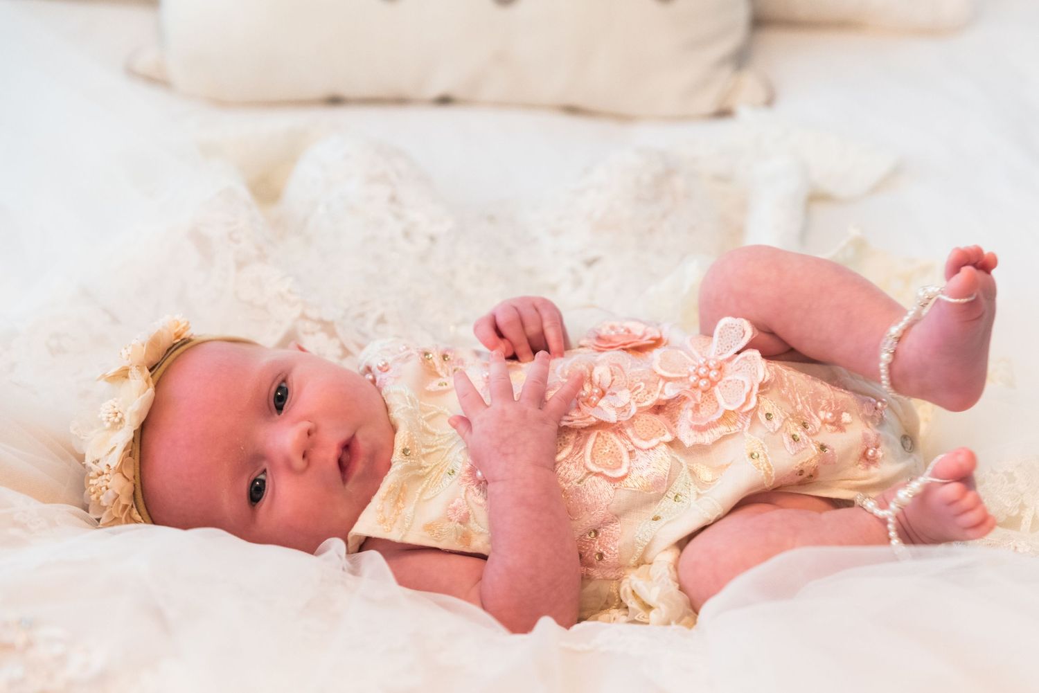 Baby wearing peach lace outfit resting on white bedding.