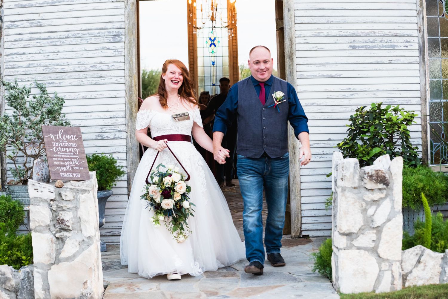 A happy couple exits a white chapel with decorative stone pillars and potted plants.