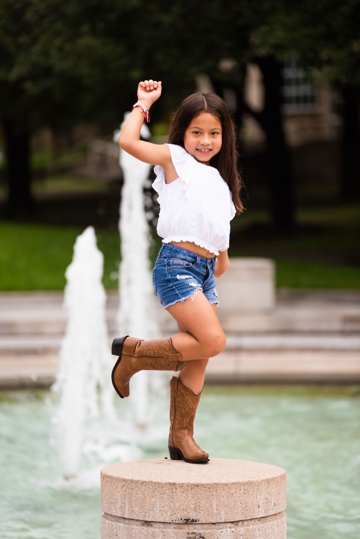 Young girl in cowboy boots strikes a playful pose by a fountain in a park setting.