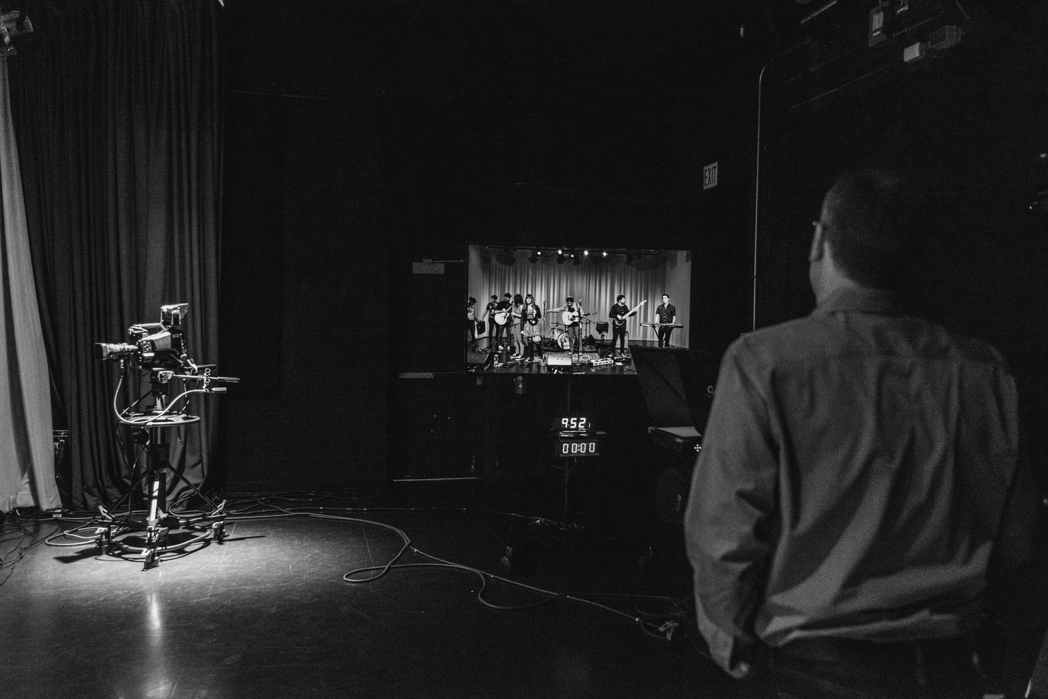 Black and white photo of empty stage with spotlight and silhouette of person watching.