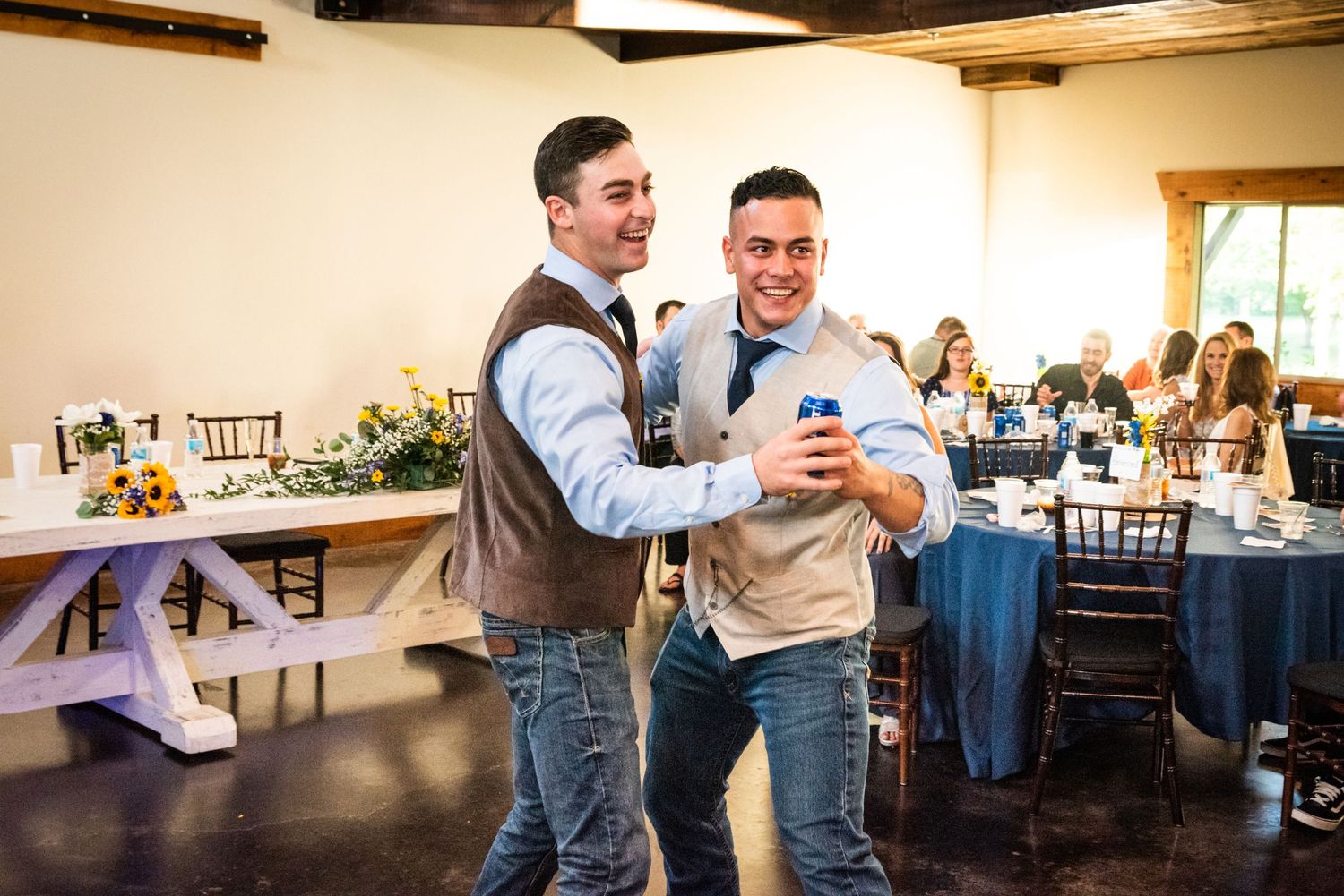 Two groomsmen in vests share a laugh while looking at a phone during the wedding reception.