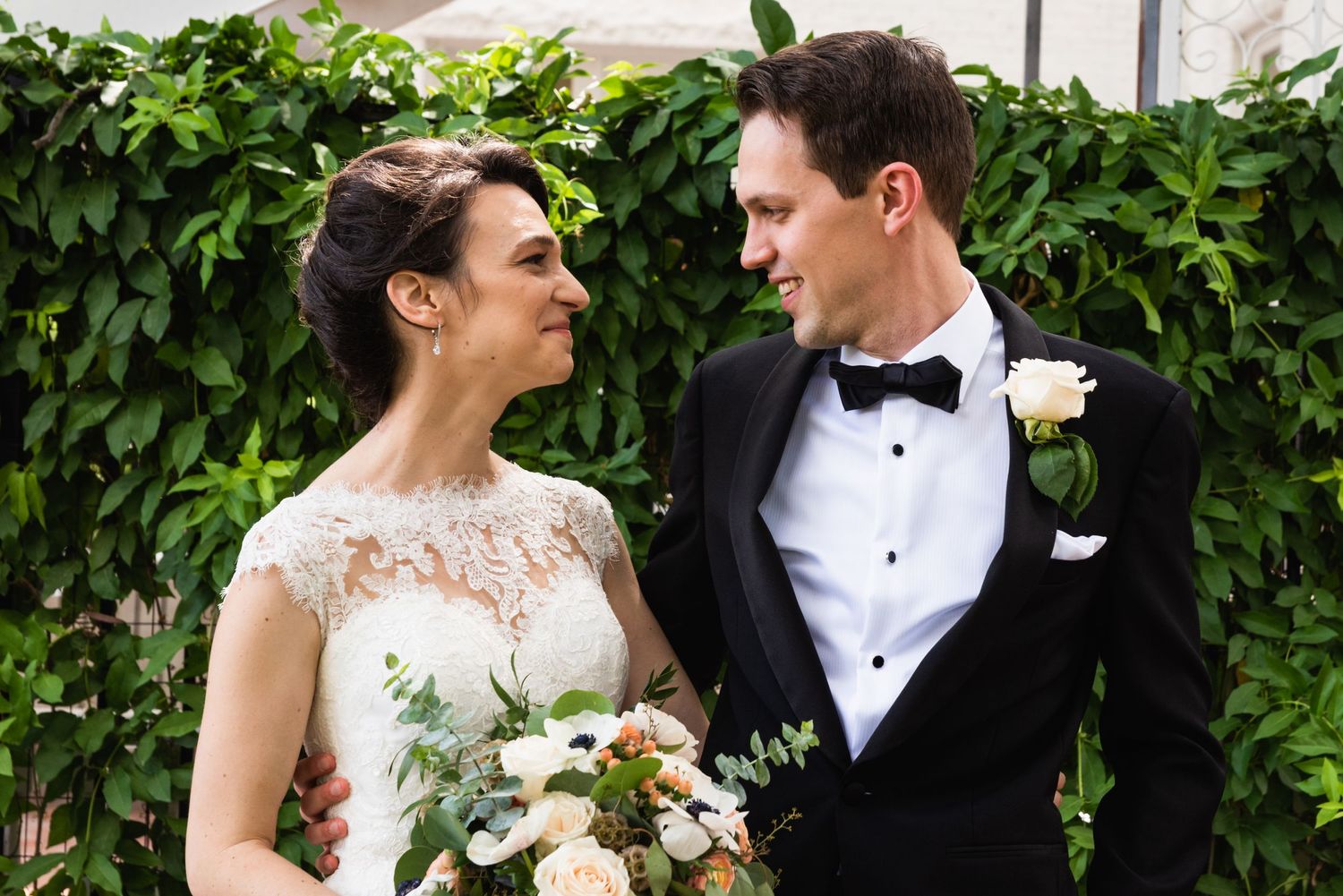 A romantic outdoor wedding portrait featuring lace details and a white bouquet against a leafy green backdrop.