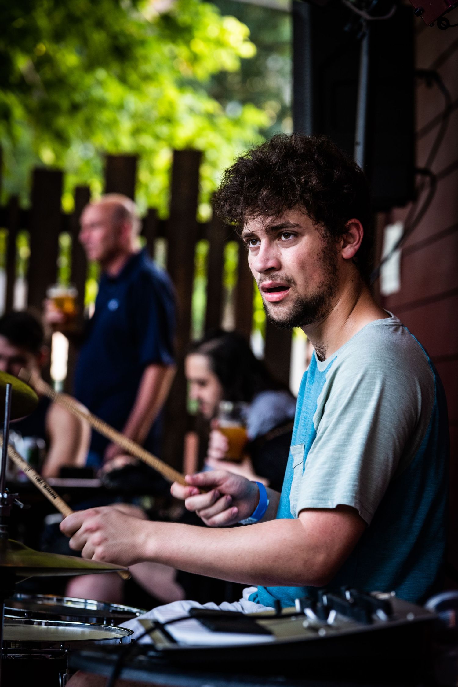 Drummer performing at outdoor venue with green foliage in background.