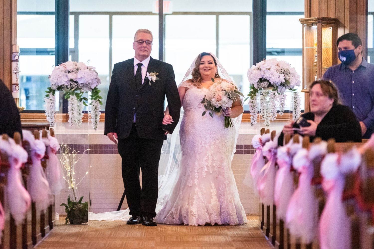 Couple walks down aisle decorated with pink floral arrangements in church setting.