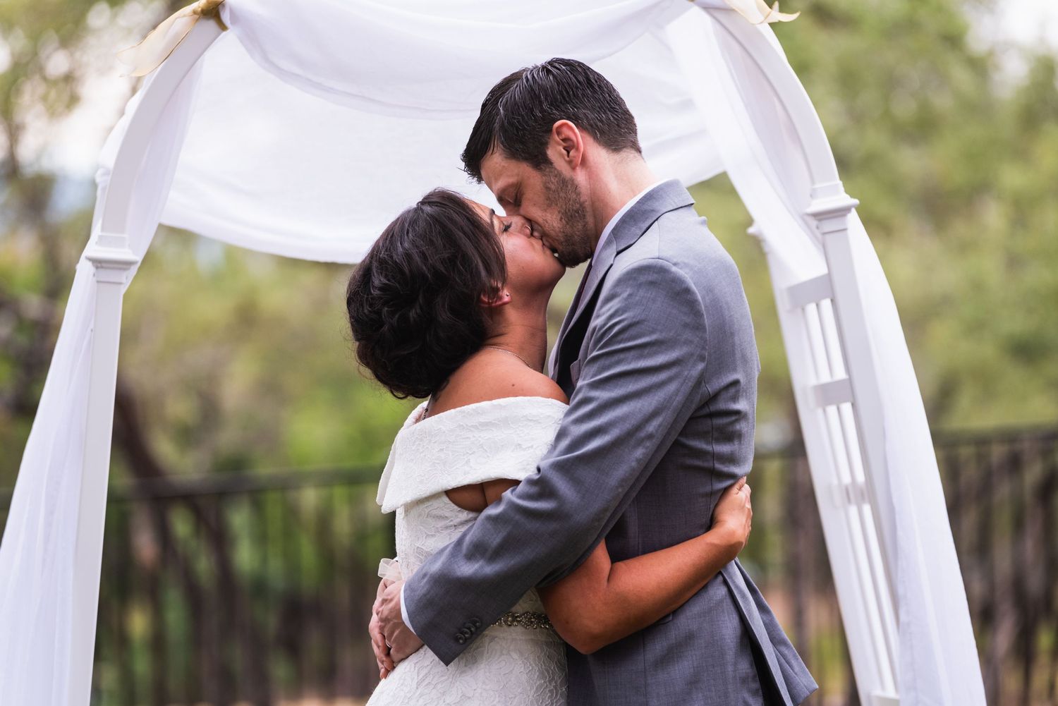 Romantic wedding kiss under white arch during outdoor garden ceremony.