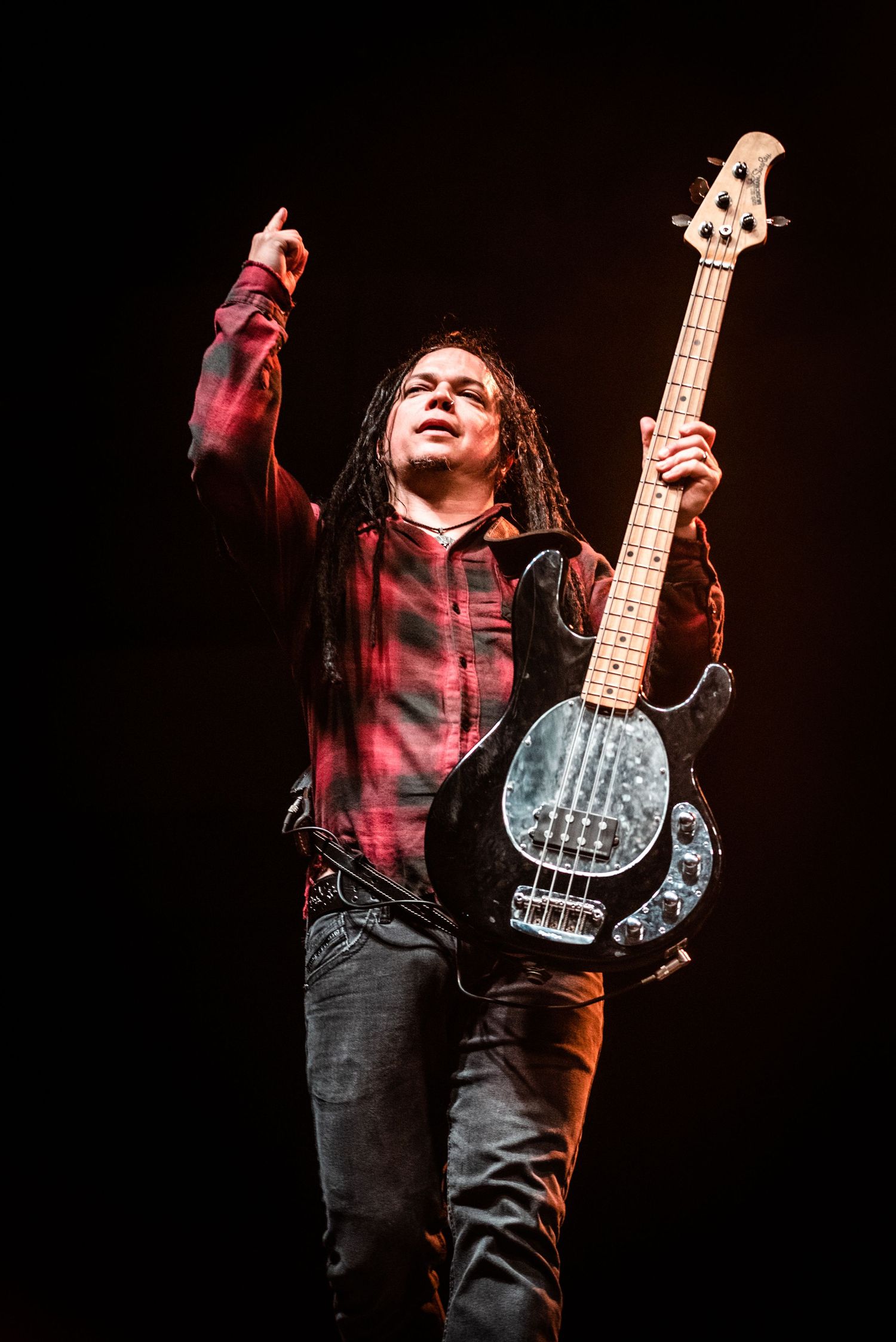 Bass guitarist in plaid shirt performs energetically on stage with raised arm.