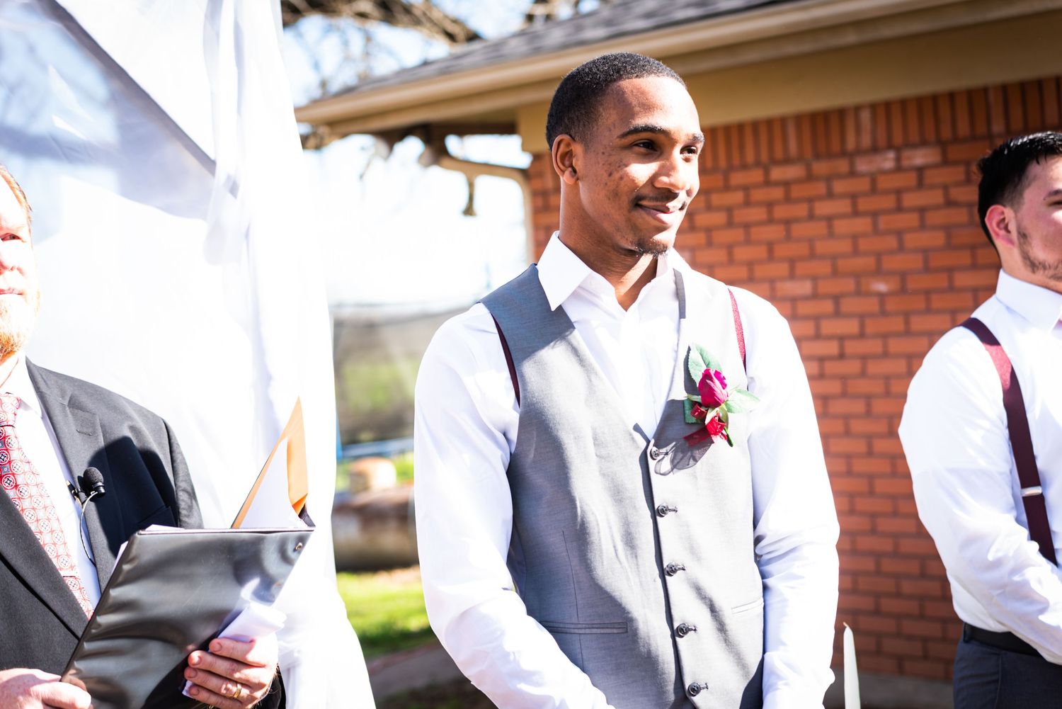 A groom in a light gray vest smiles during an outdoor wedding ceremony.
