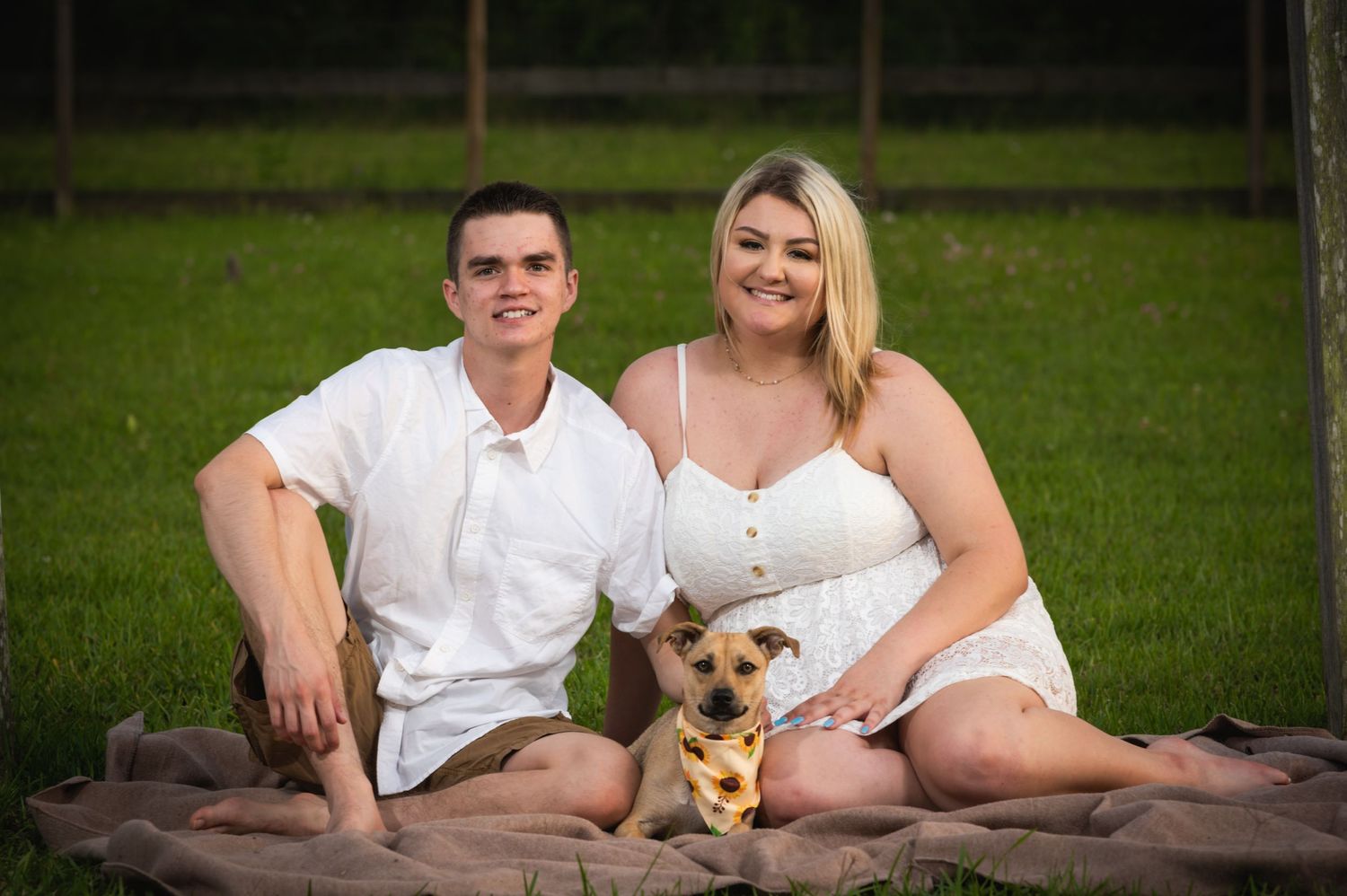 A couple poses with their small dog on a brown blanket in a grassy field.