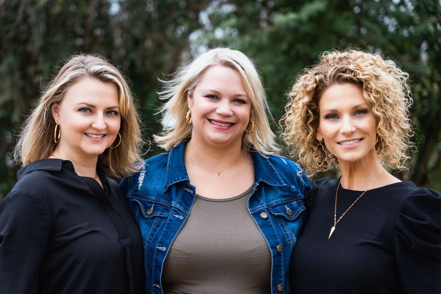 Three women in casual attire pose together for an outdoor portrait.