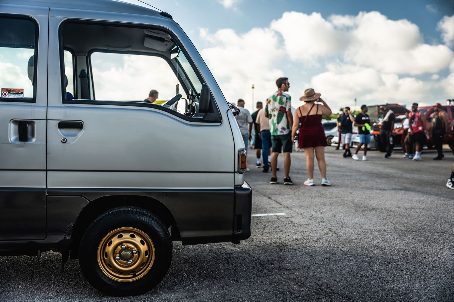 Silver van parked at a crowded outdoor car meet with spectators in background.