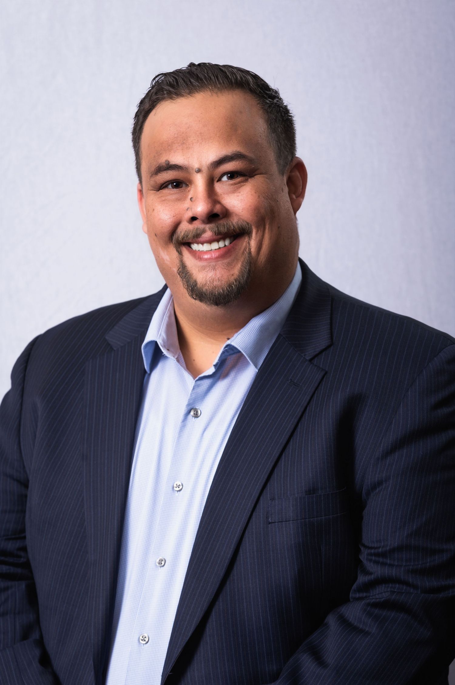 Professional headshot of someone in a navy pinstripe suit and light blue shirt against a gray background.