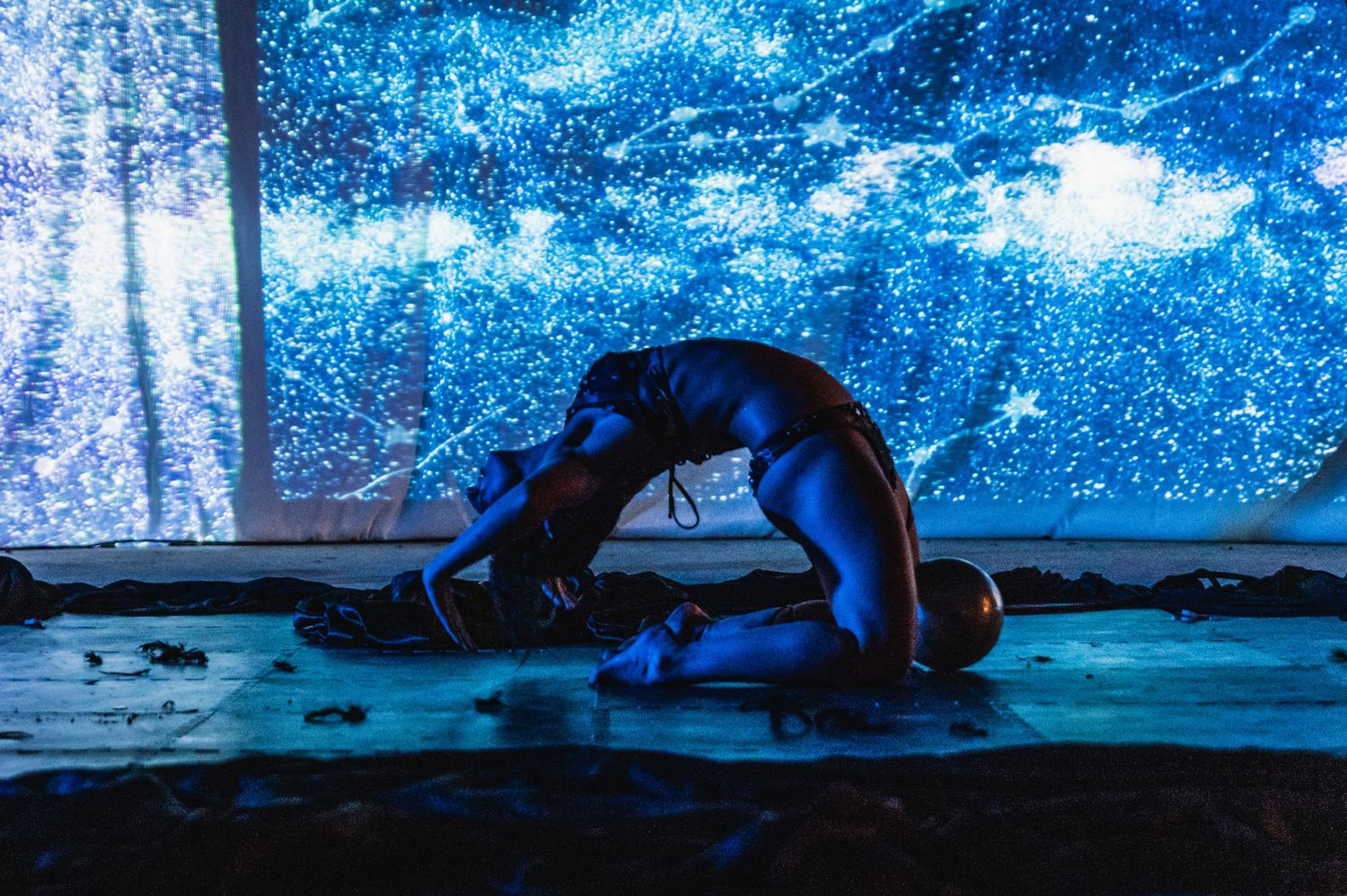 Silhouetted performer in backbend pose against cosmic blue projection background.