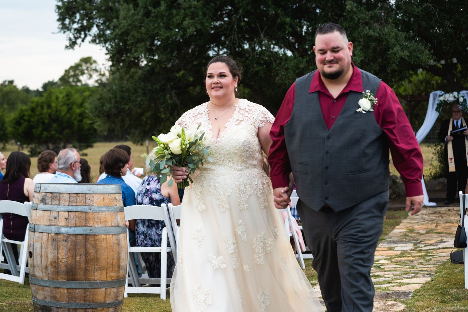 Newlyweds walking past rustic wine barrel after outdoor wedding ceremony.