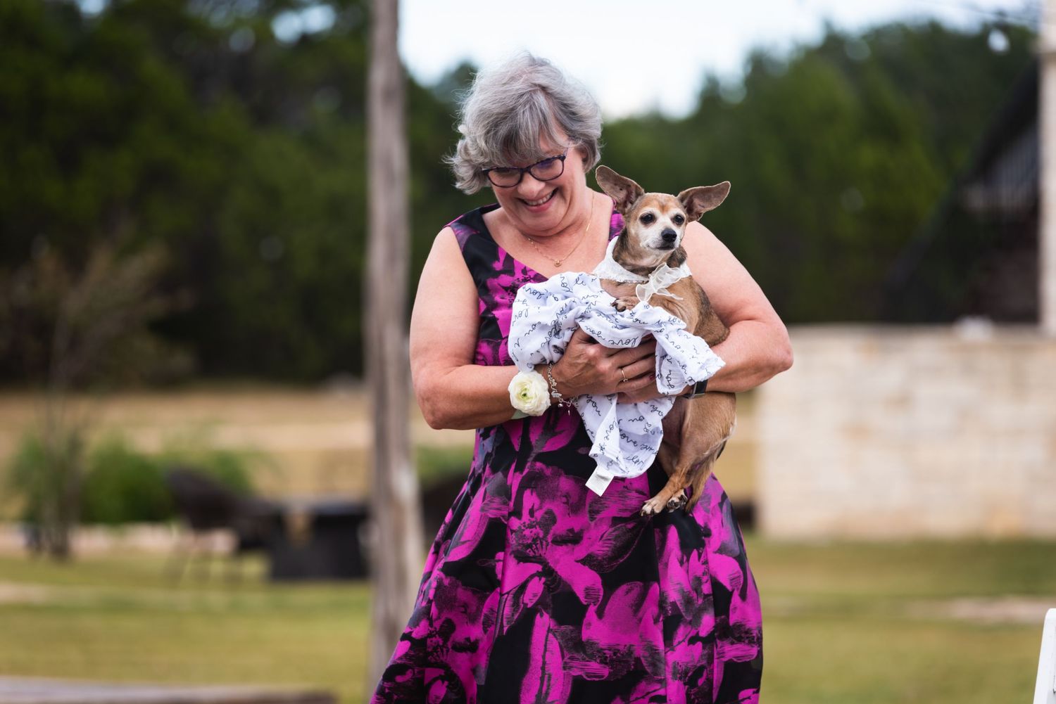 Someone wearing a vibrant pink and black dress holds a small white dog outdoors.