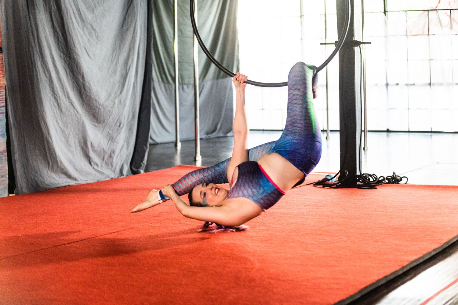 Aerial artist in blue outfit performs floor routine on red mat in studio space.
