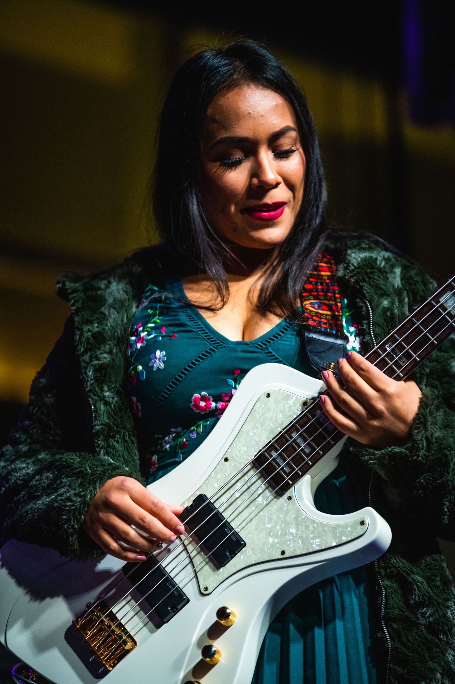 Close-up of guitarist playing white electric bass while wearing floral dress and black fuzzy jacket on stage.