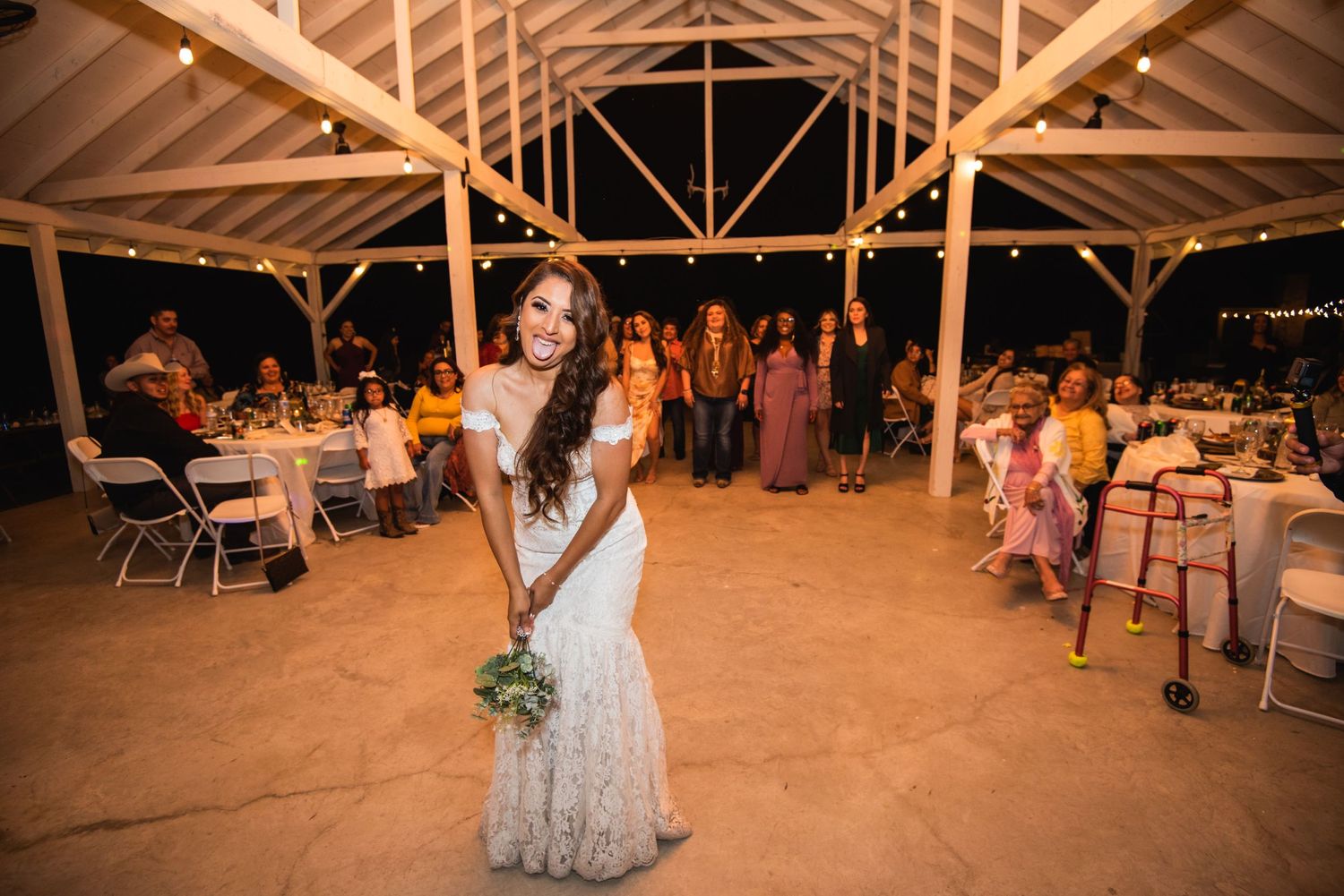 A wedding reception celebration in a rustic barn venue with guests seated at tables and string lights overhead.