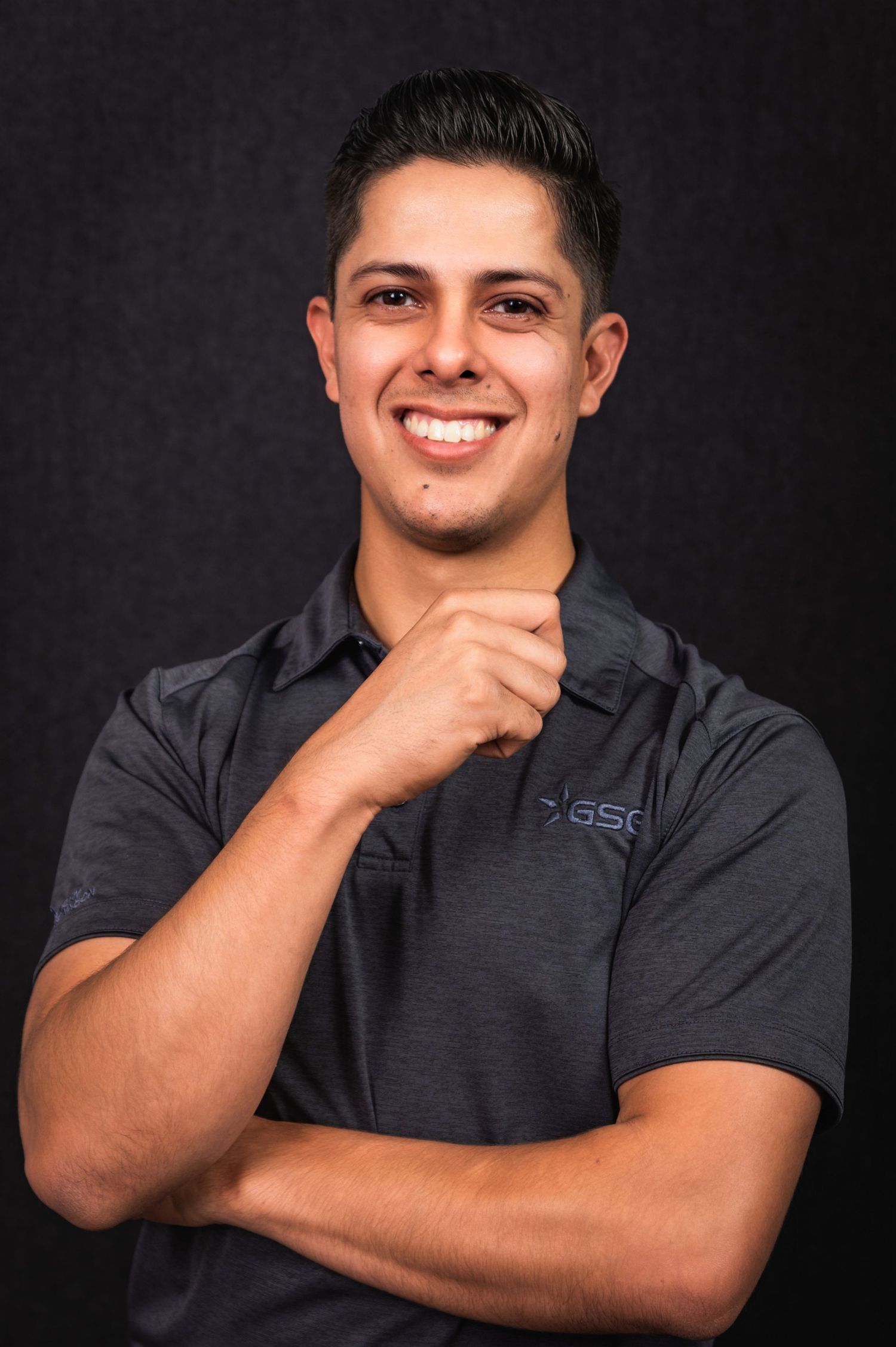 Corporate portrait of man in dark polo with hand on chin against black backdrop.