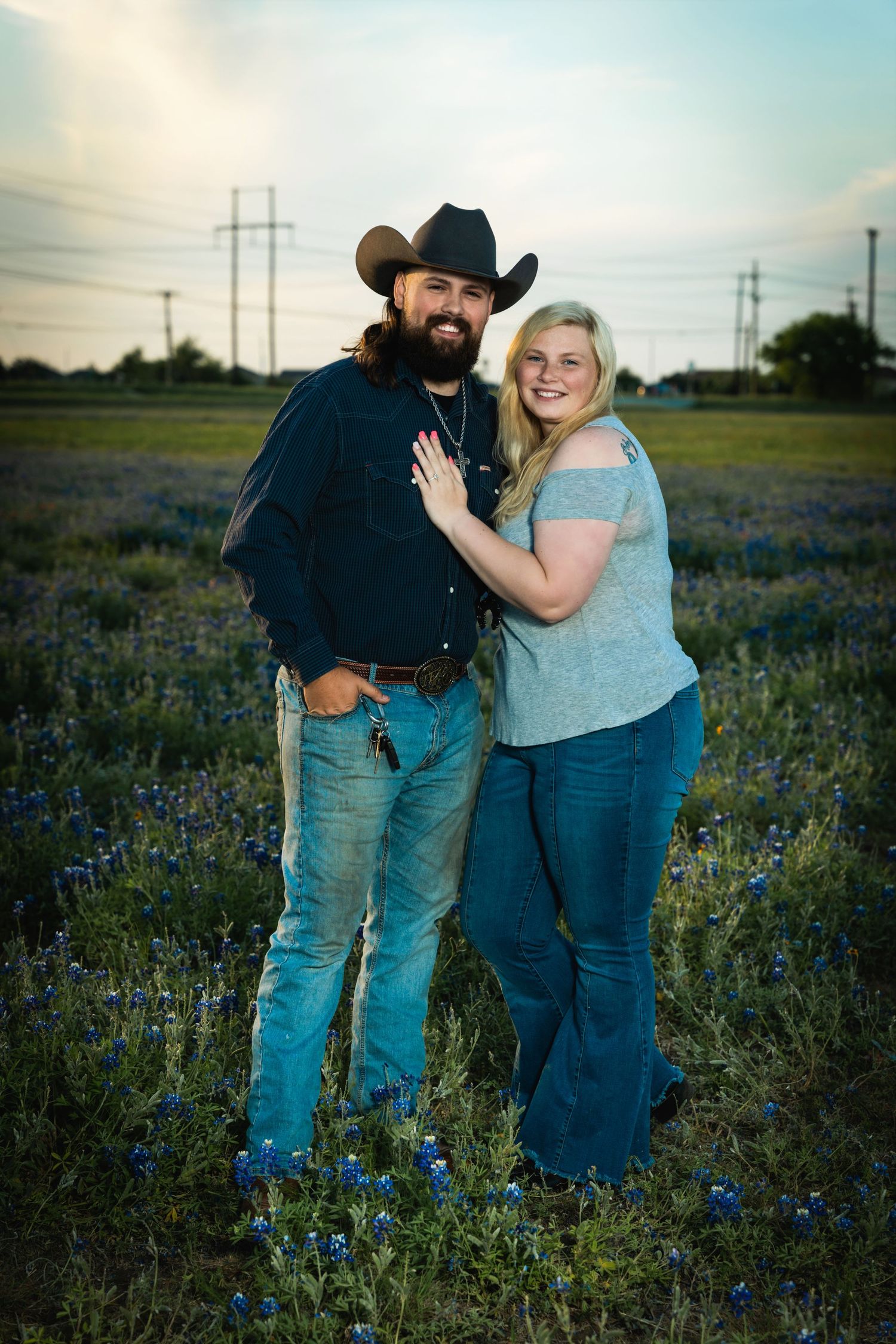 A couple poses in a field of bluebonnets at sunset with power lines in the background.