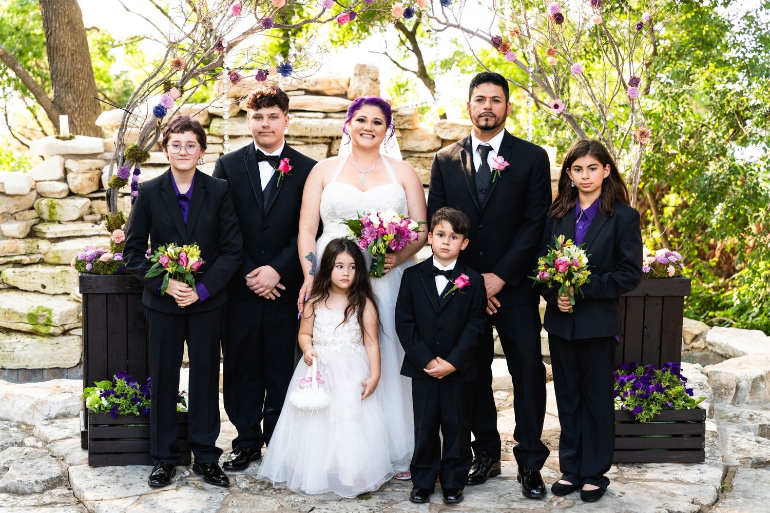 Formal wedding party portrait with black and purple attire against stone wall backdrop.