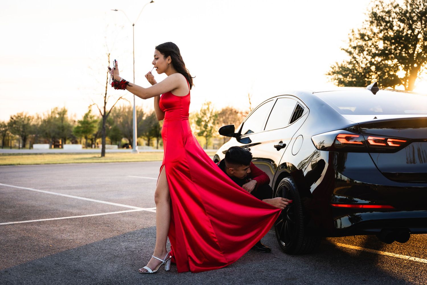 Dramatic portrait of a flowing red dress against a black luxury car at sunset.