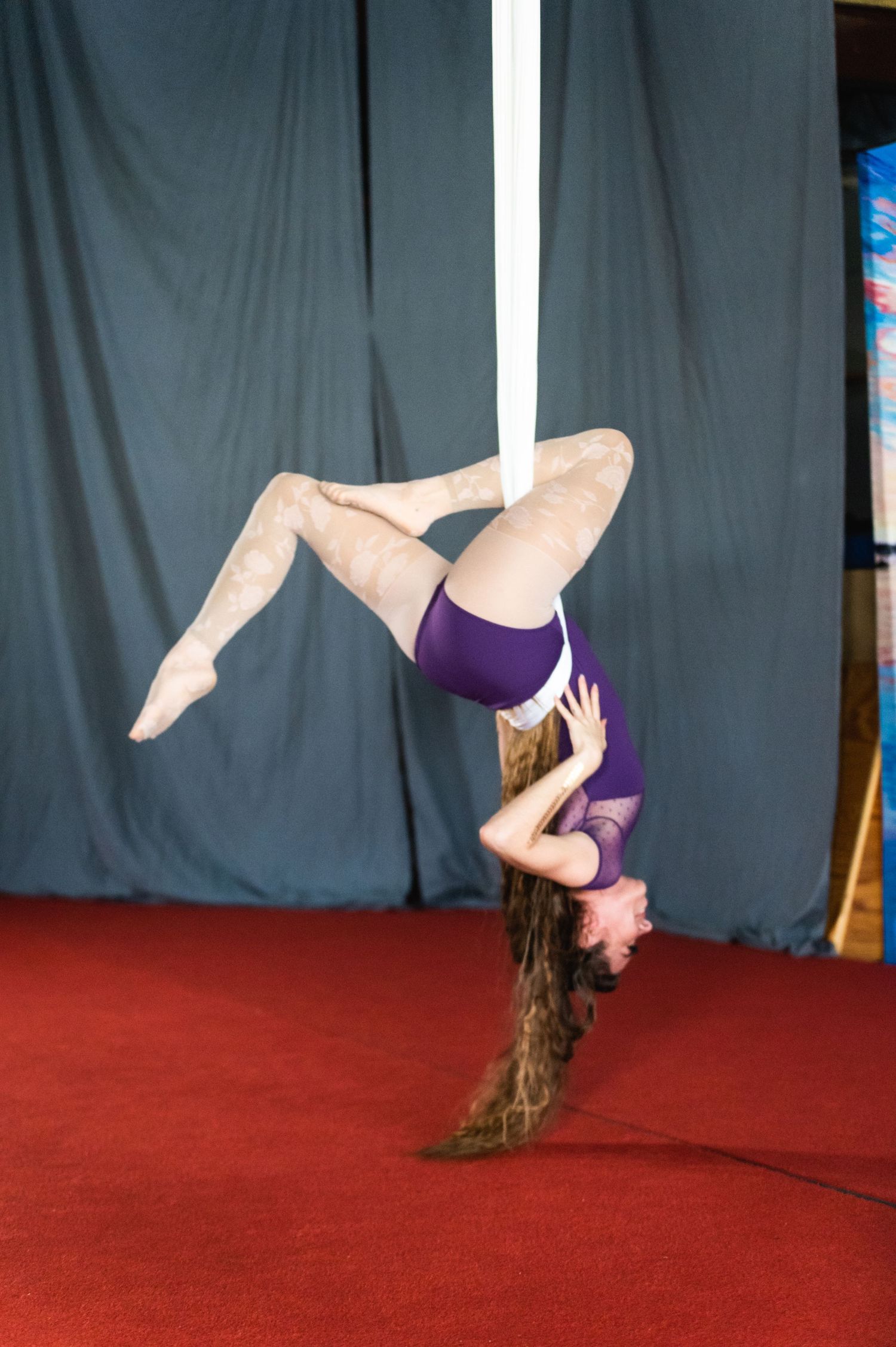 Aerial artist in purple leotard performs inverted pose on white silks against dark background.
