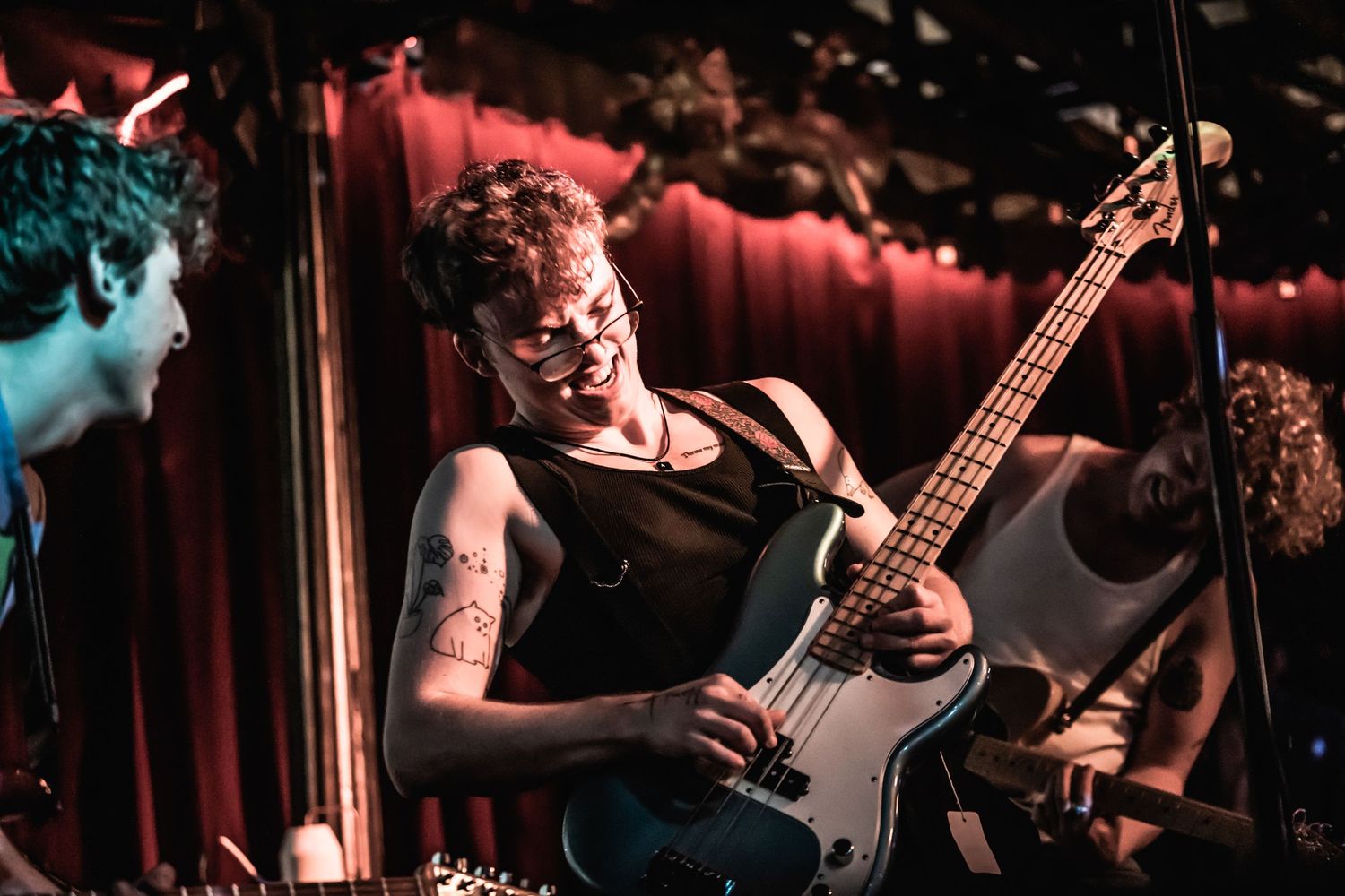 A musician performs passionately on bass guitar at a dimly lit venue with red stage lighting.