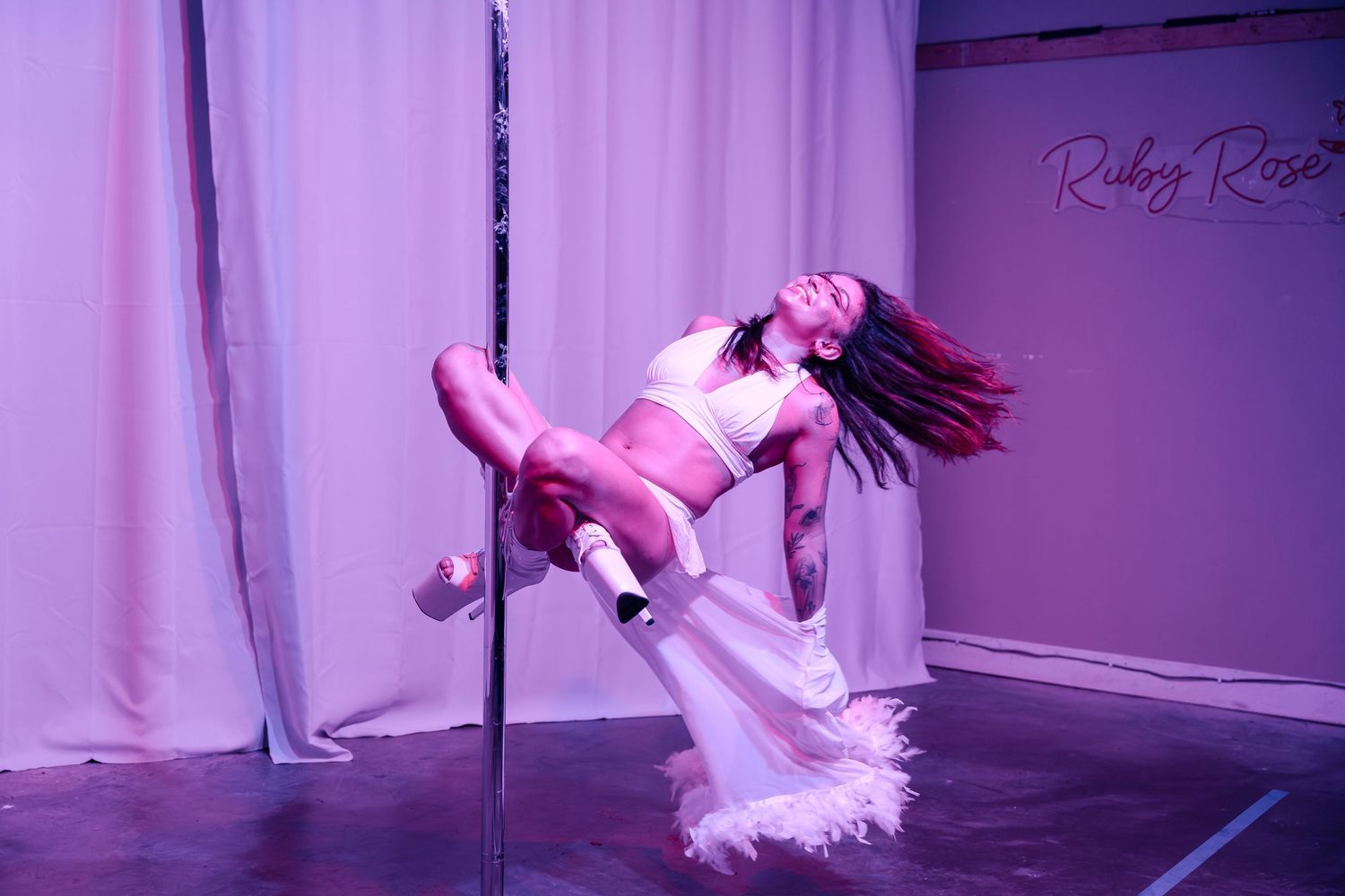Pole dancer performs an artistic move in pink lighting at a studio performance space.