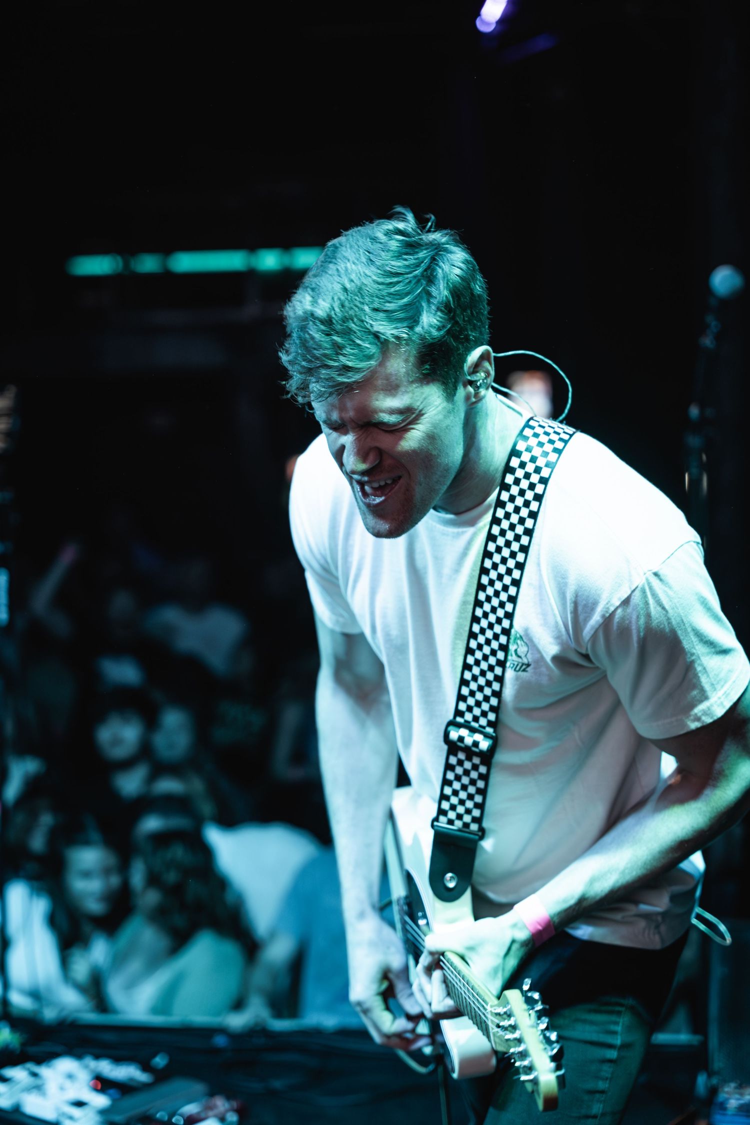 Guitarist in white shirt with checkered strap plays passionately under blue lighting.