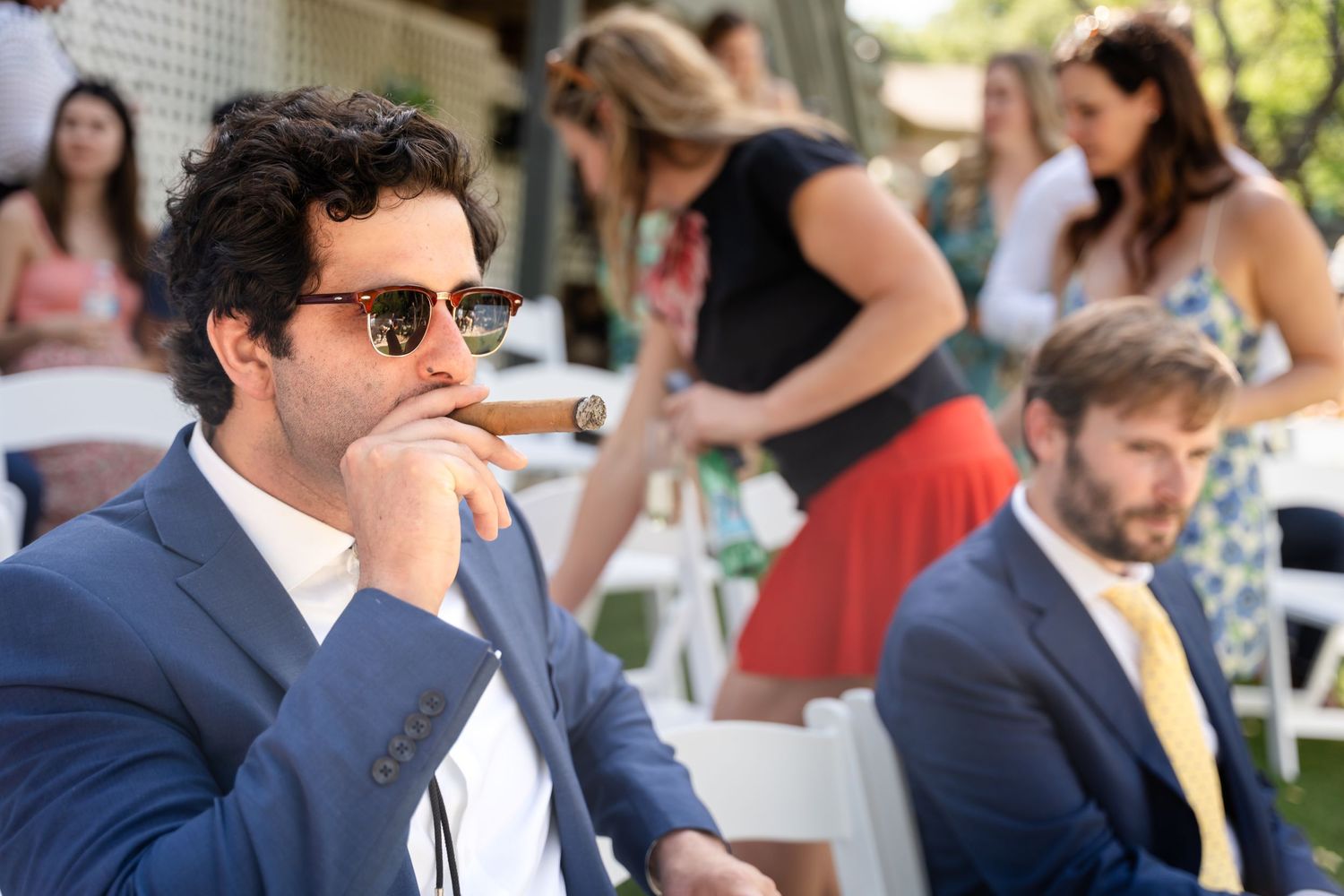 A guest enjoying a cigar at an outdoor event while wearing sunglasses and a navy suit.
