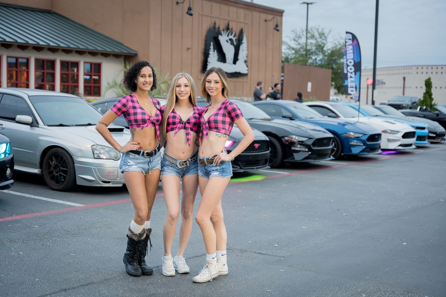 Three women in coordinated pink plaid outfits pose at a car meet.