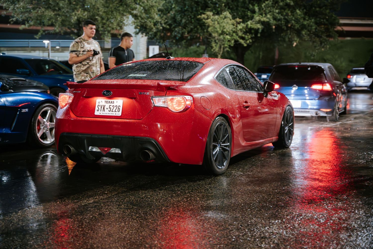 Red sports car with aftermarket modifications on wet pavement at a car show.
