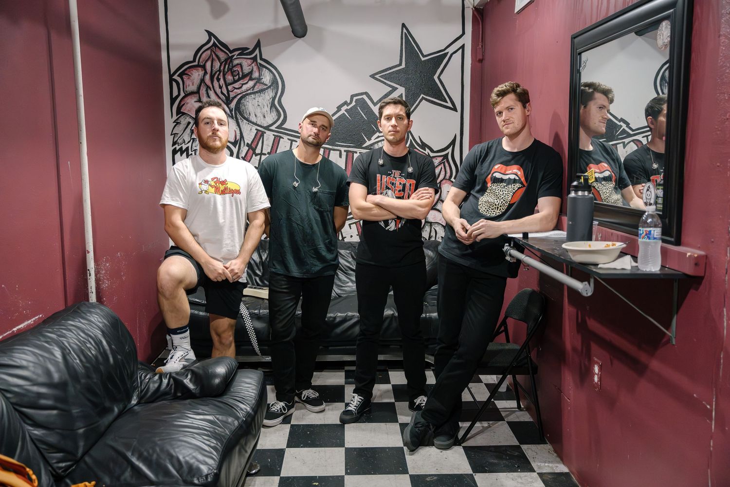 A group poses together in a checkered-floor room with band posters and decor on the walls.