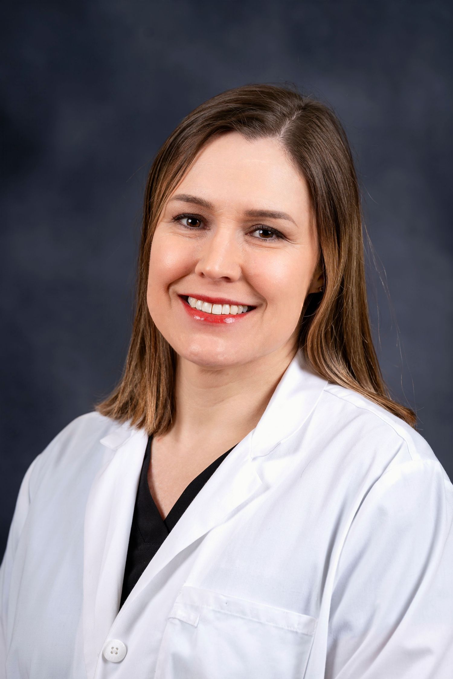Healthcare professional in white lab coat and dark shirt smiling in front of dark background.