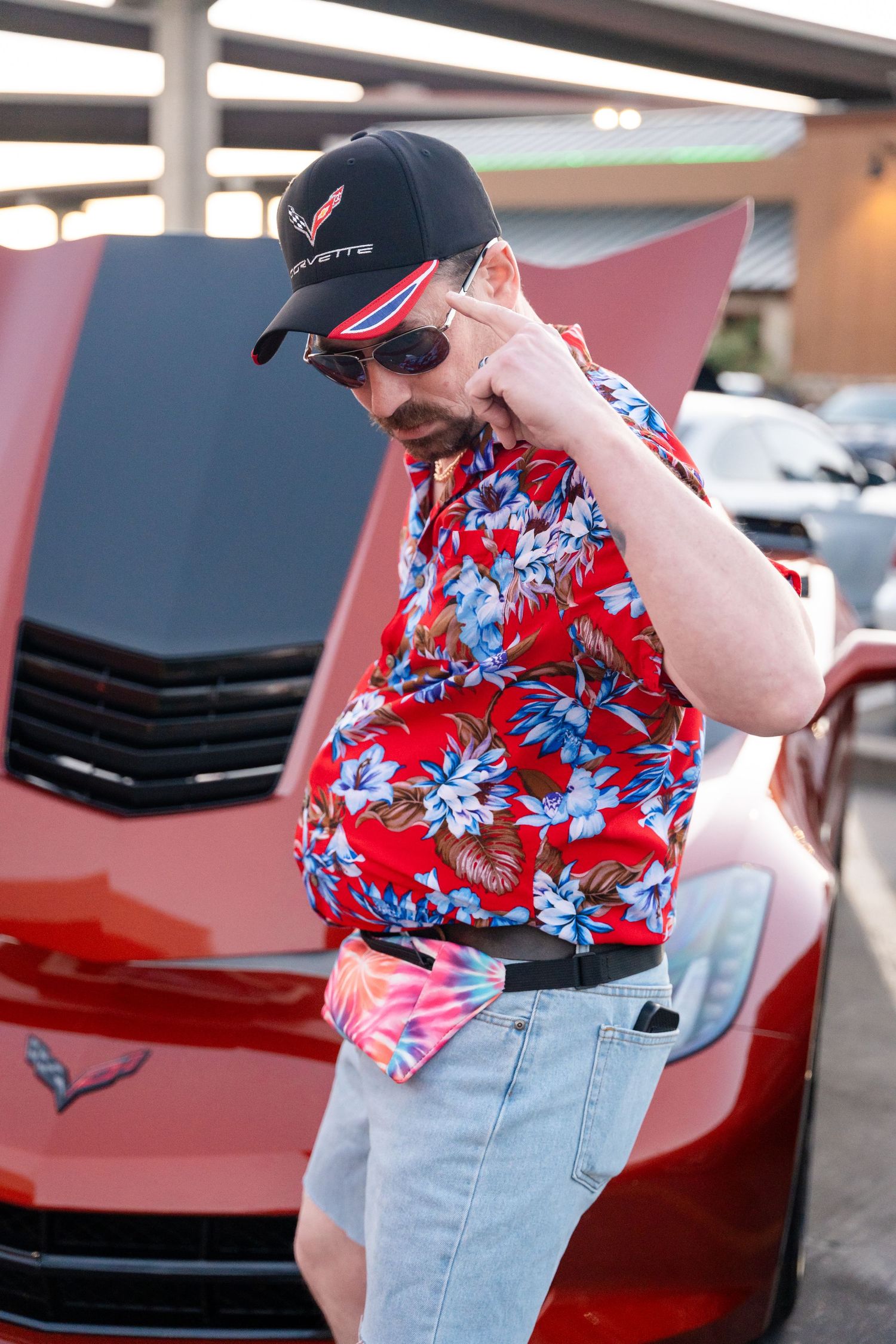 Person in red floral shirt and baseball cap posing next to a red sports car.