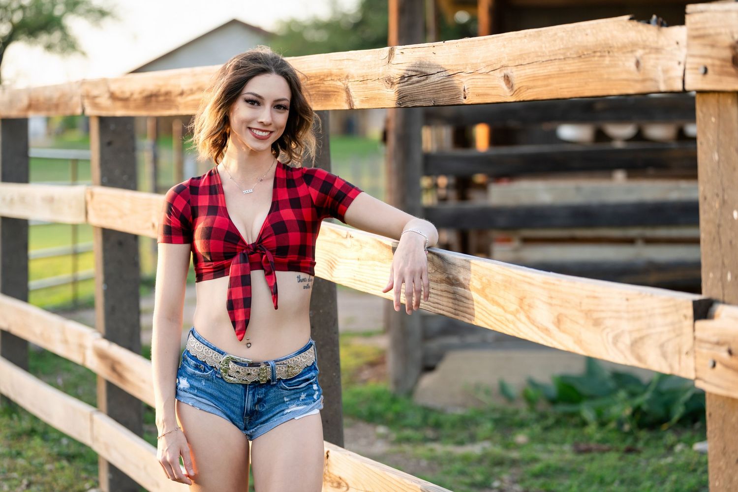 Western-inspired outfit with red plaid top and denim shorts posed by wooden ranch fence.