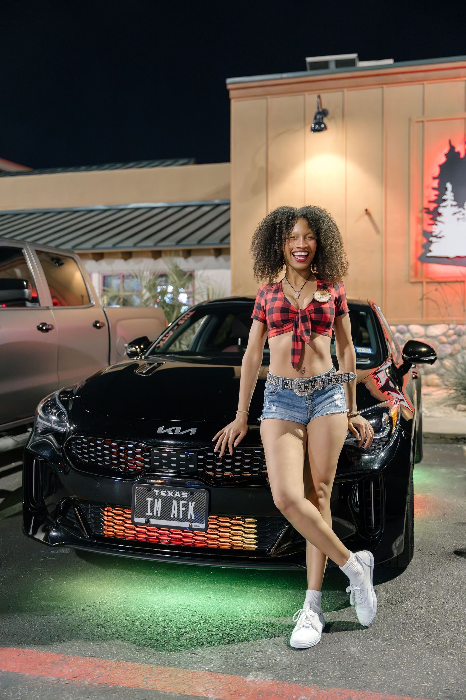 Woman in red plaid top and denim shorts posing by black sports car with custom lighting.