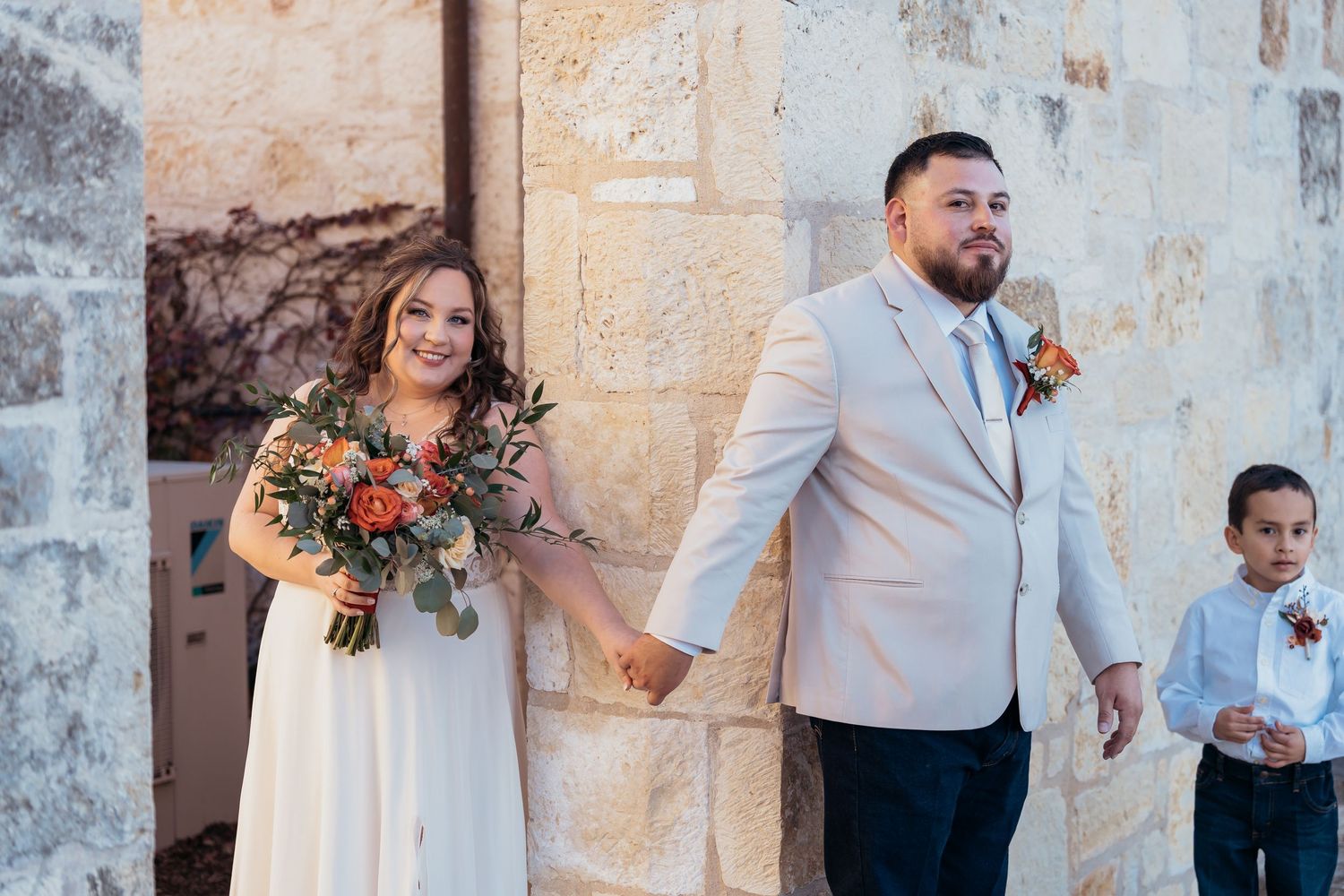 Couple and child posing outside rustic stone building during wedding celebration.