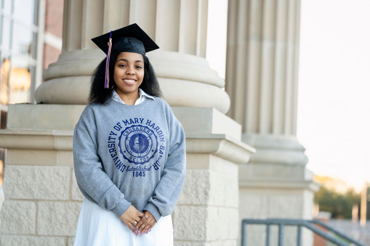 A graduate in a University of Mary Hardin Baylor sweatshirt poses in front of classical columns.