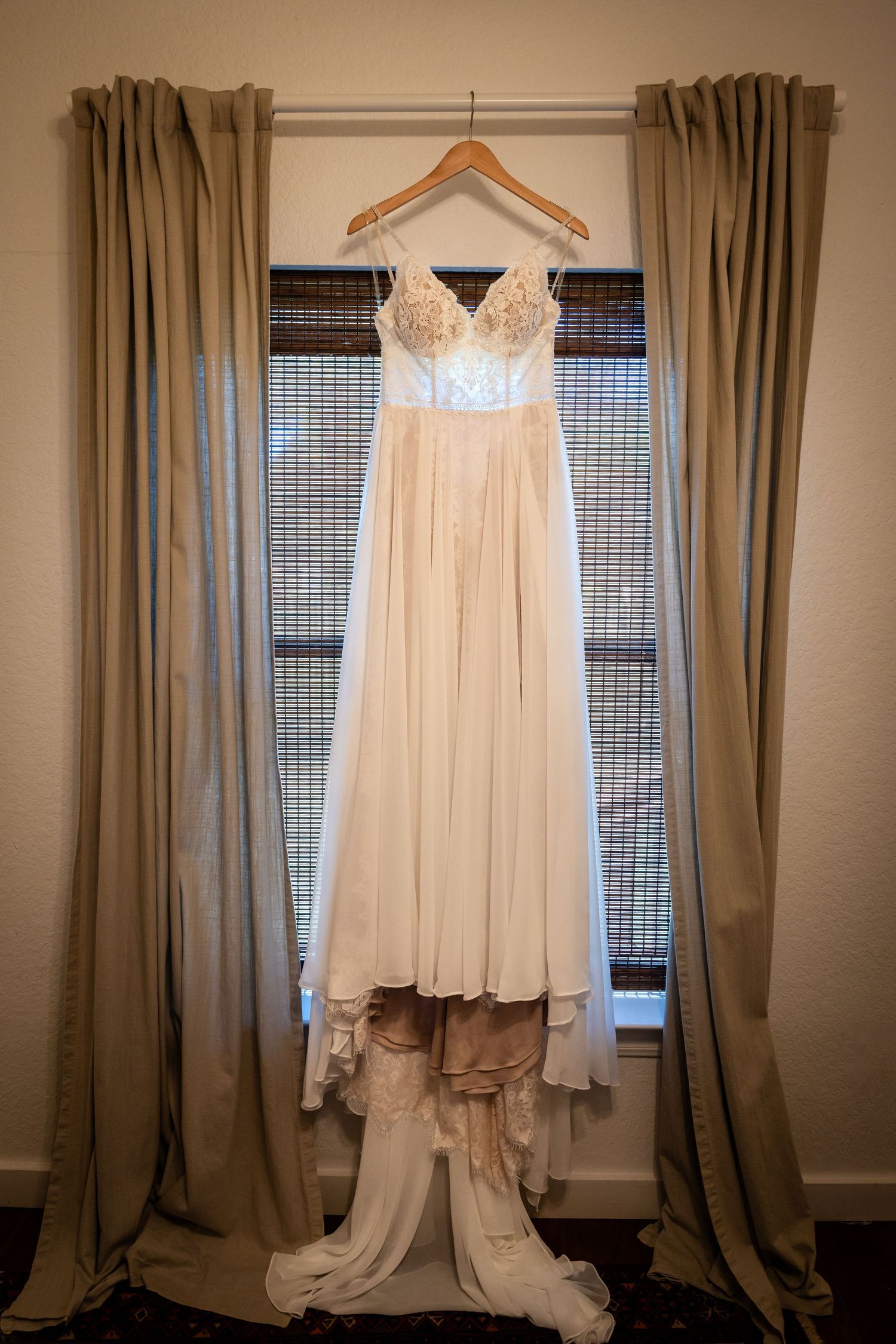 White lace wedding dress hanging between tan curtains in front of bamboo blinds.
