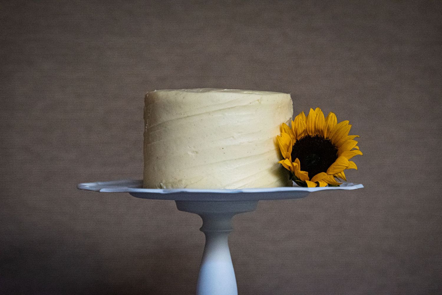 Simple white wedding cake decorated with a single sunflower on a gray pedestal stand.