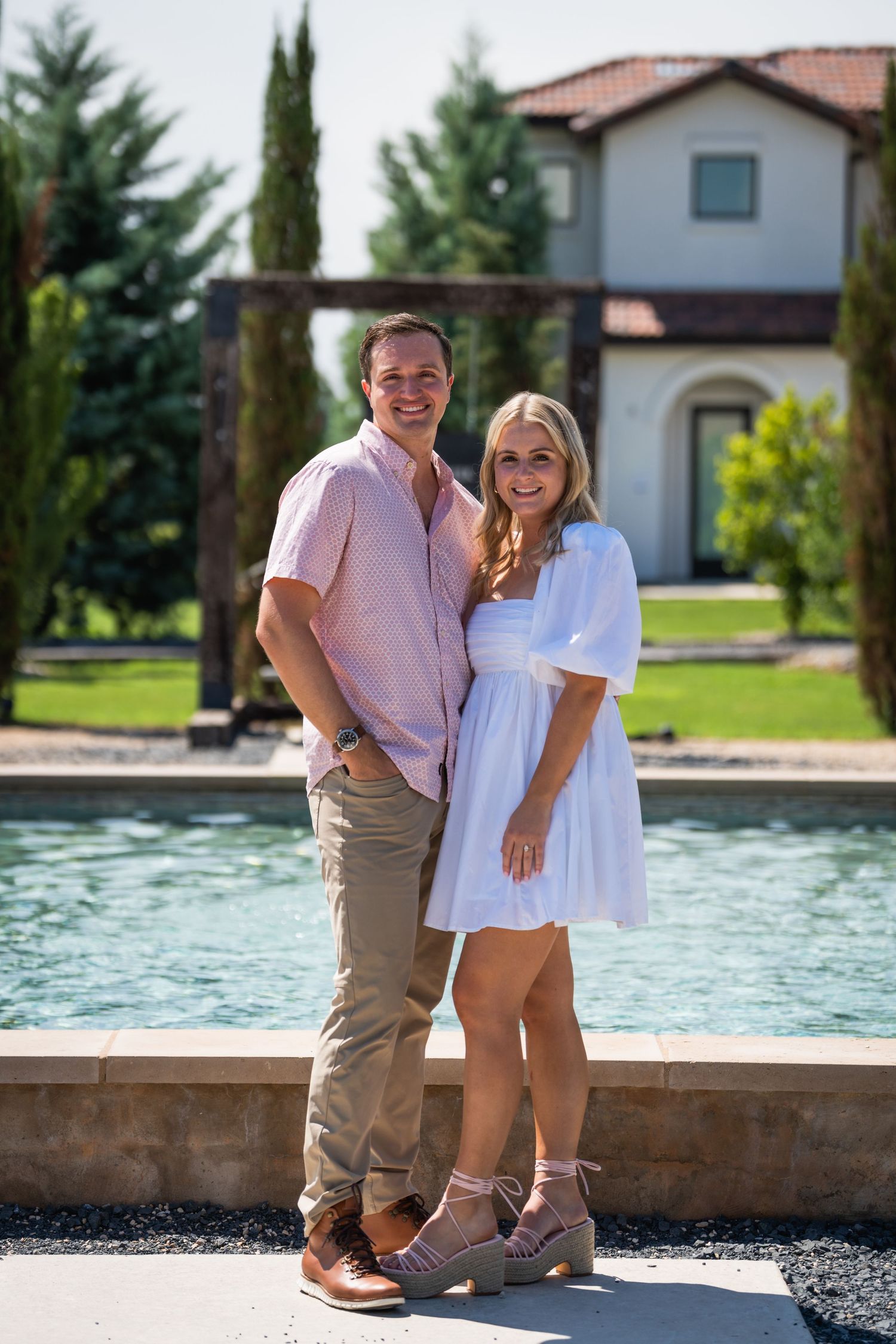 A couple poses together in summer attire next to a Mediterranean-style pool and villa.