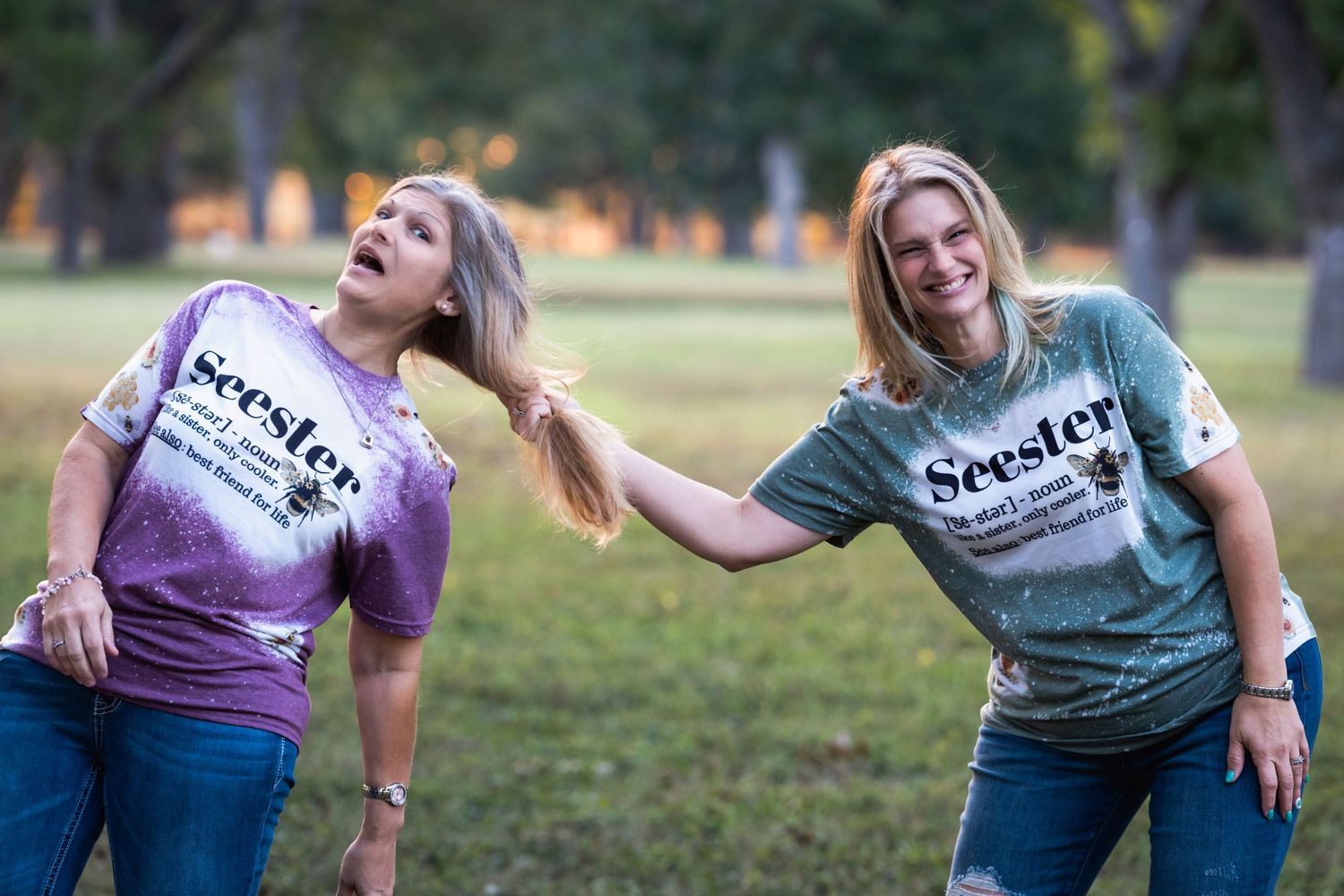 Two people in tie-dye shirts playfully pose together in a park setting at sunset.