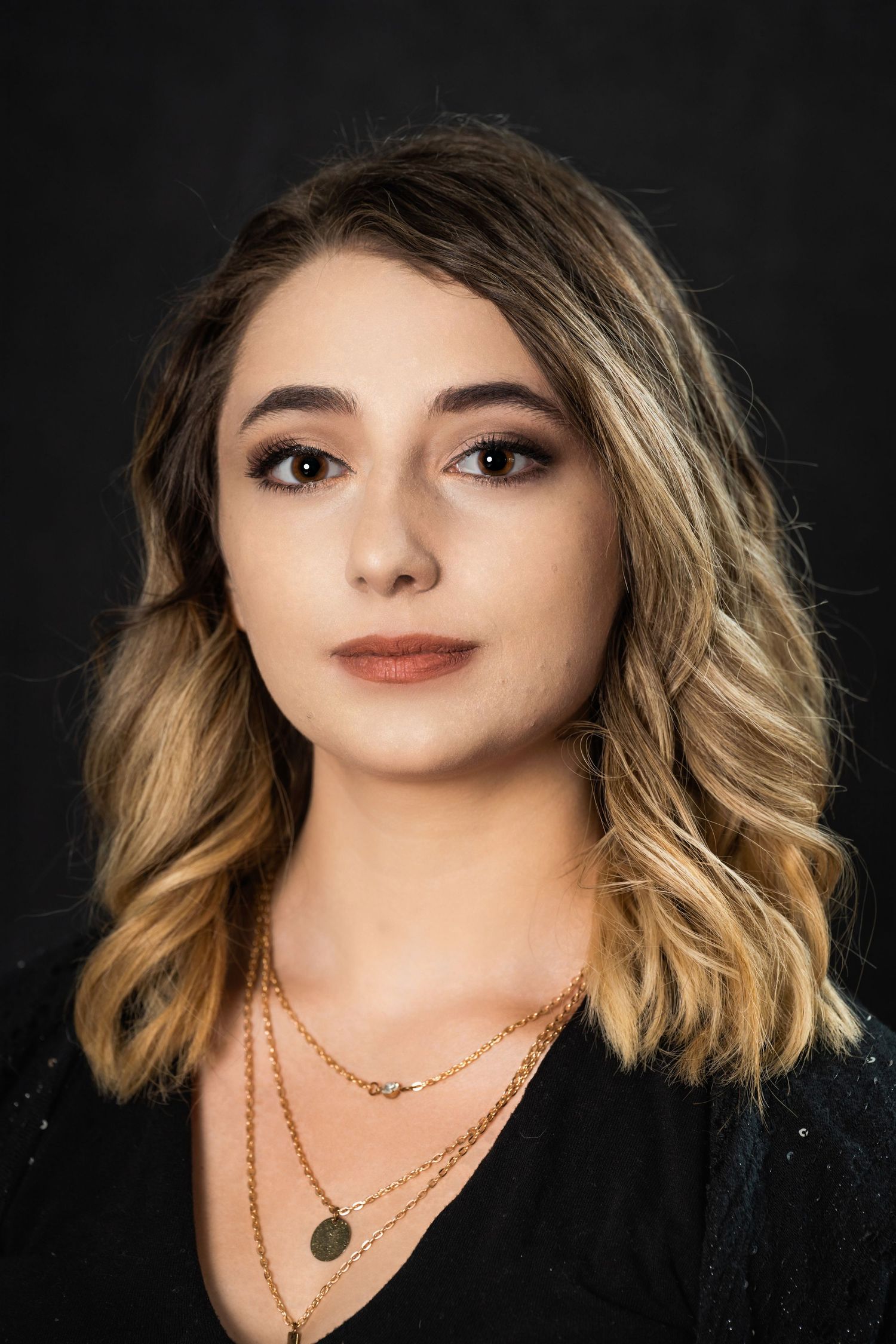 Studio portrait featuring wavy ombre hair and delicate gold necklaces on black background.