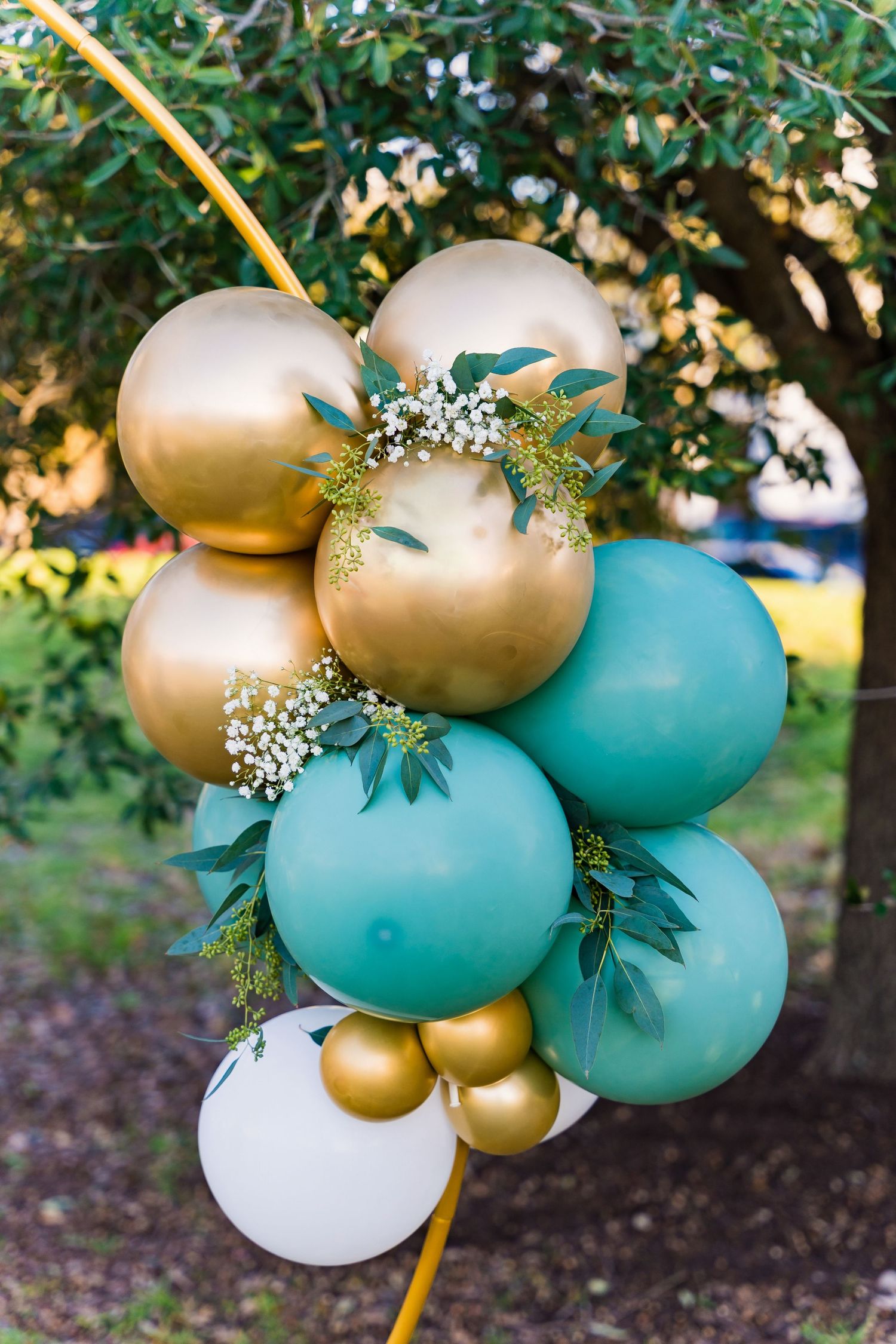 Close-up of balloon arrangement featuring gold, turquoise and white balloons with baby's breath and green foliage.